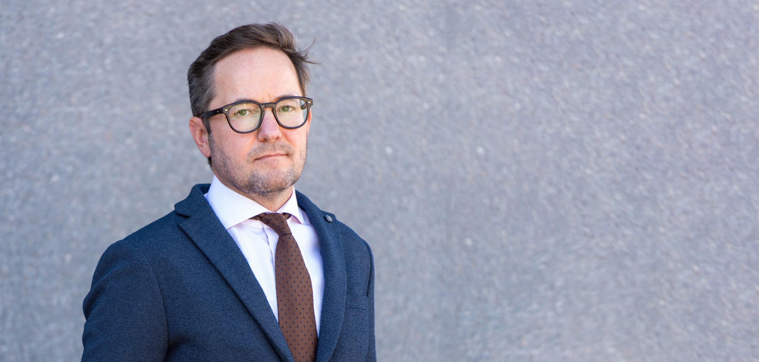 En mann i mørk dress med hvit skjorte og brun slips, står foran en grå vegg.
