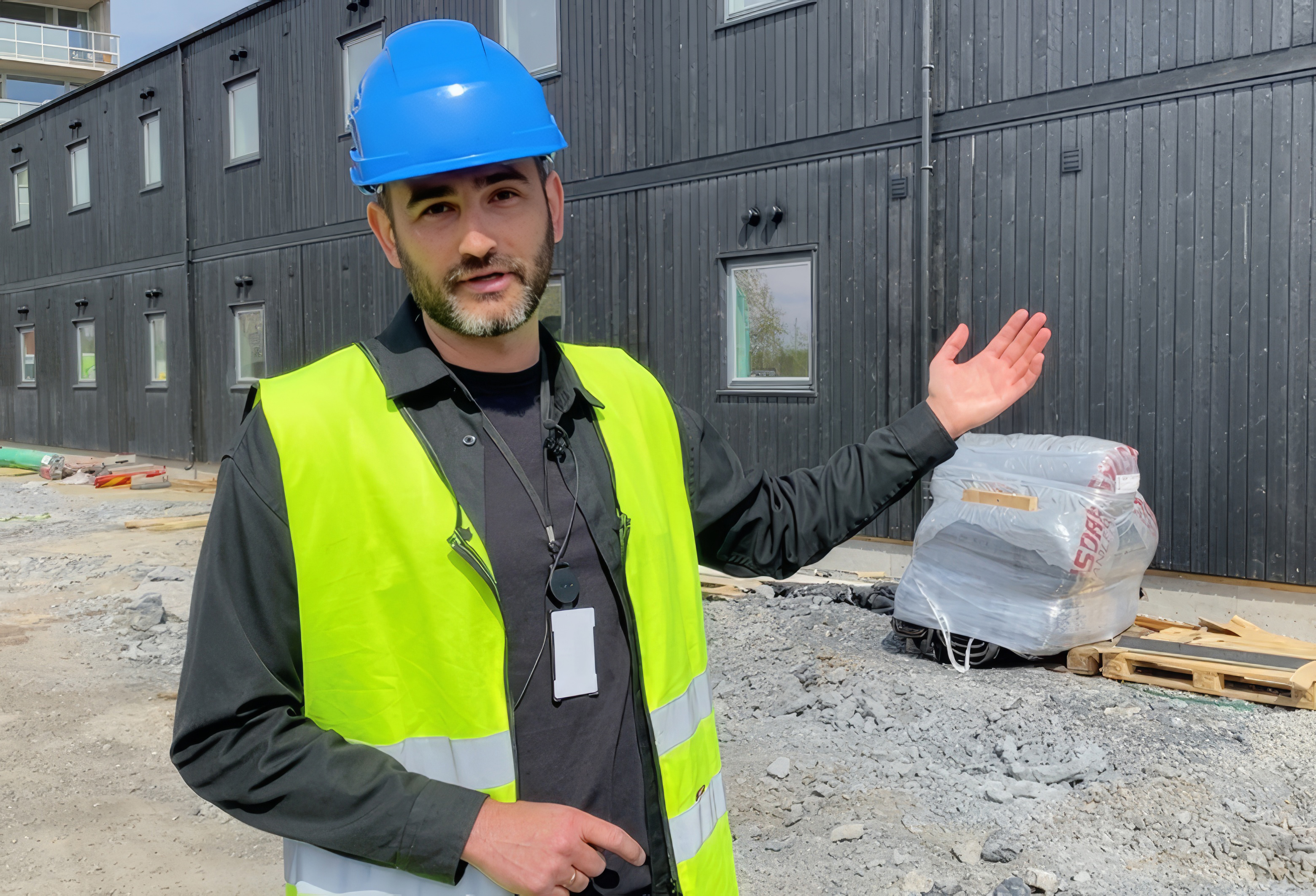 Mann, med blå hjelm og svart skjorte, står foran svarte brakker på en arbeidsplass.
