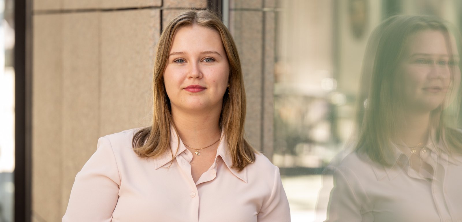 Ung kvinne med lyst hår står foran en murvegg og et vindu; hun ser rolig mot kameraet, og refleksjonen vises i glasset.