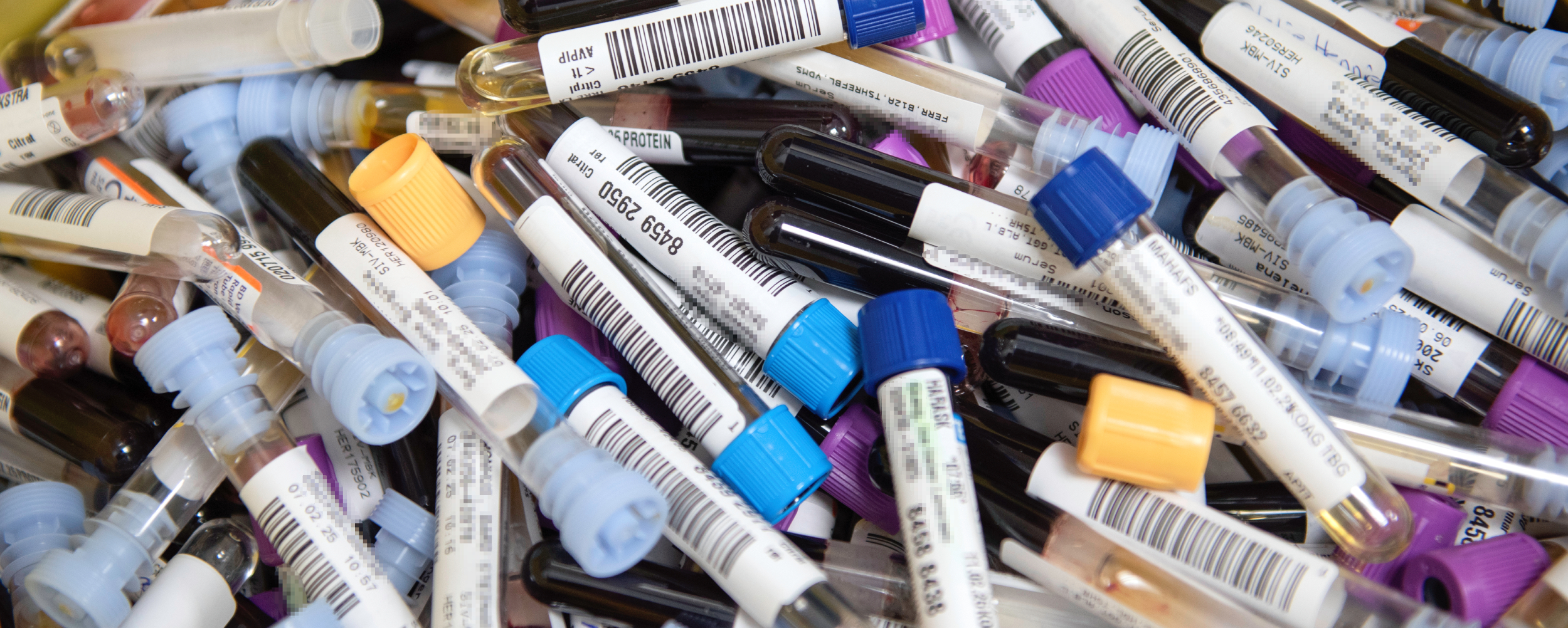 Bildet viser en samling med blodprøverør med ulike fargede korker, hver med en etikett og strekkode for identifikasjon