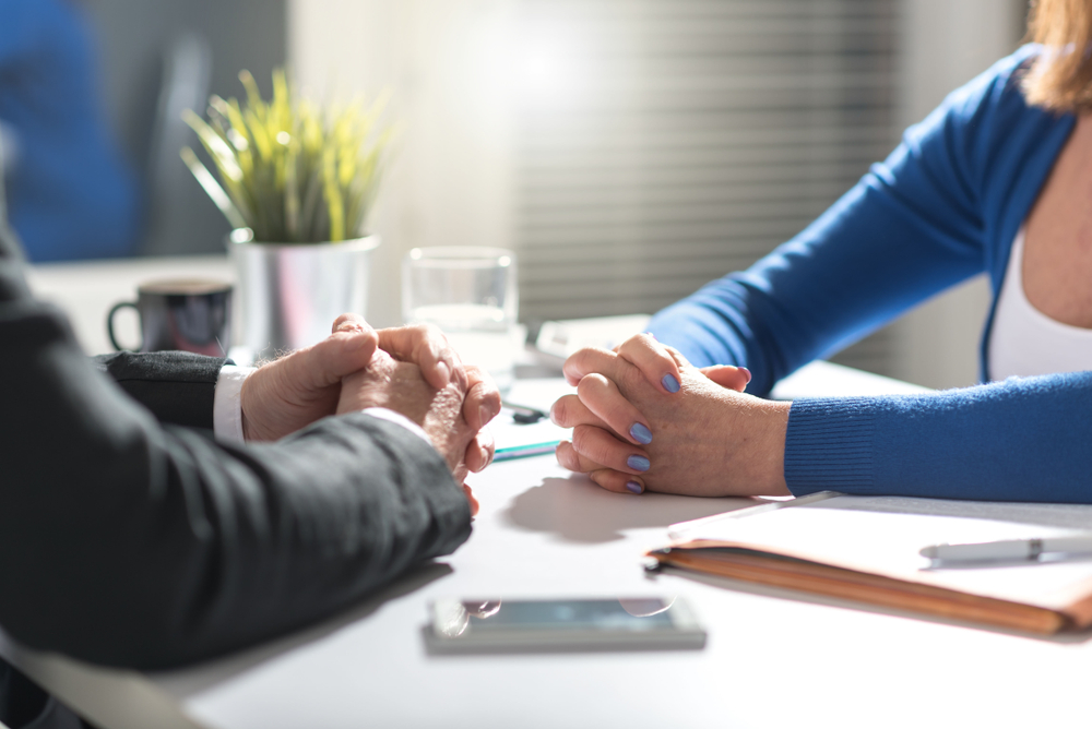 Two people are sitting at a table with their hands folded, in conversation in an office.