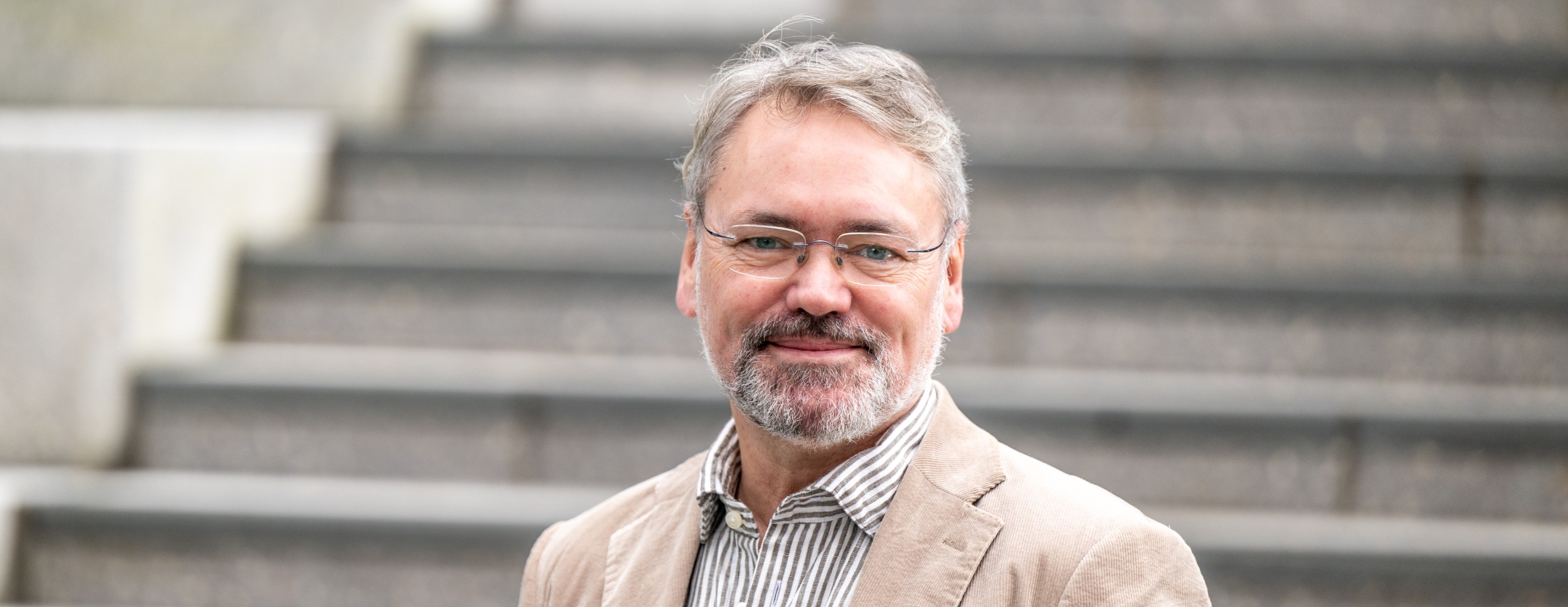 En smilende mann med briller og grått skjegg står foran en trapp, iført beige blazer og stripete skjorte. Han utstråler ro og trygghet.