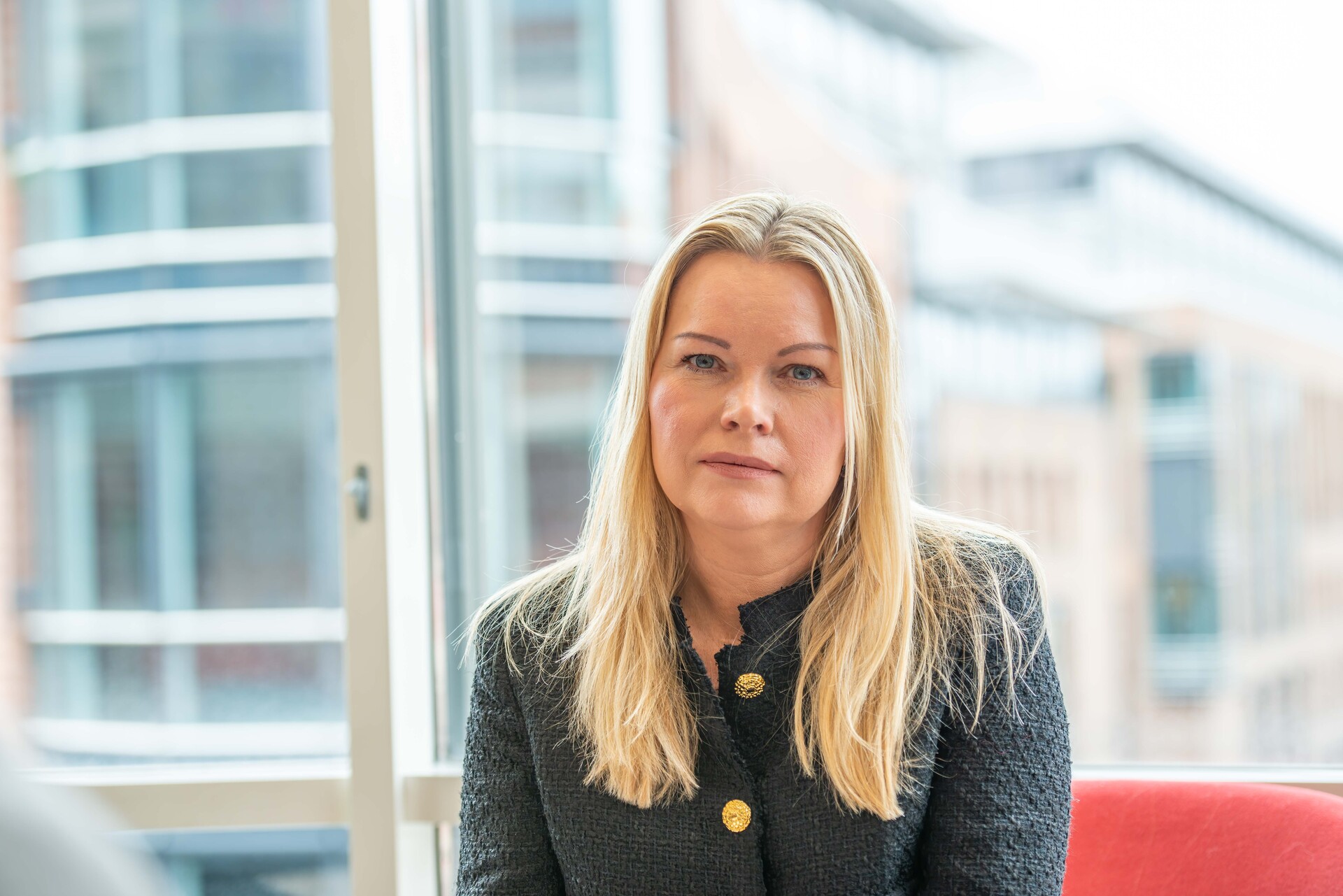 Blonde kvinne sitter i en rød stol foran store vinduer. Har på seg en grønn jakke med store gullknapper