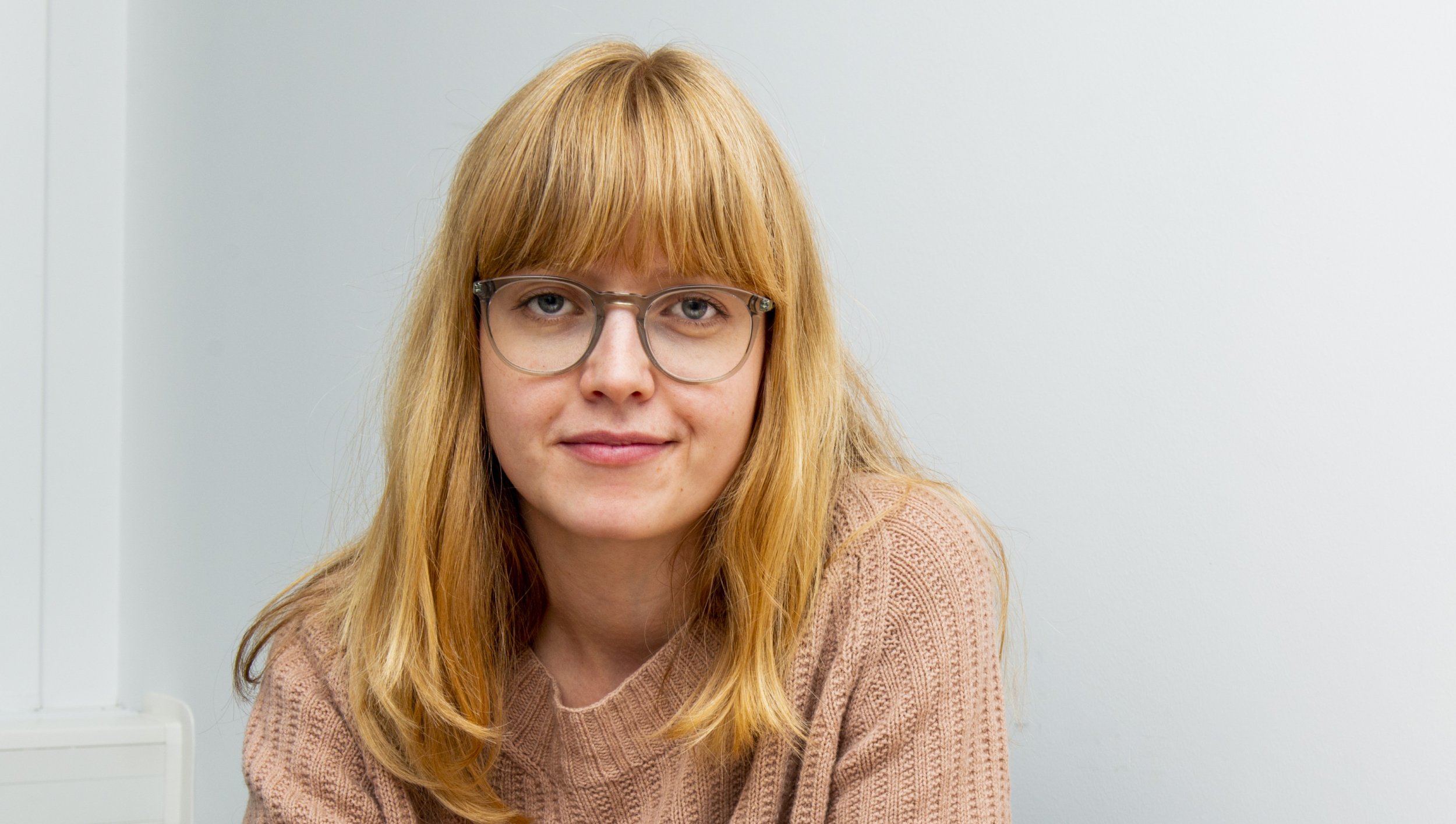 En kvinne med langt, blondt hår og briller ser rett mot kameraet. Hun har på seg en beige strikkegenser.