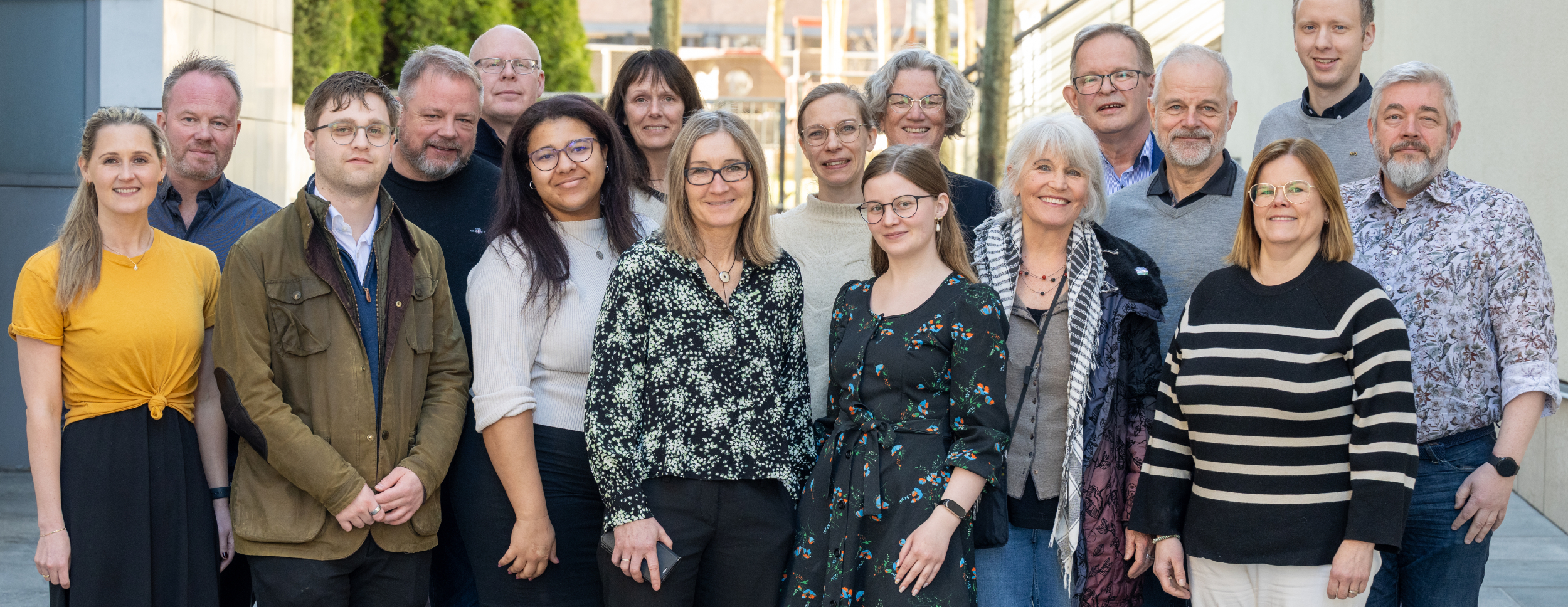 Bildet viser 18 voksne kvinner og menn som står ute i dagslys og smiler mot kamera. De er uformelt kledd, med bygninger og trær i bakgrunnen.