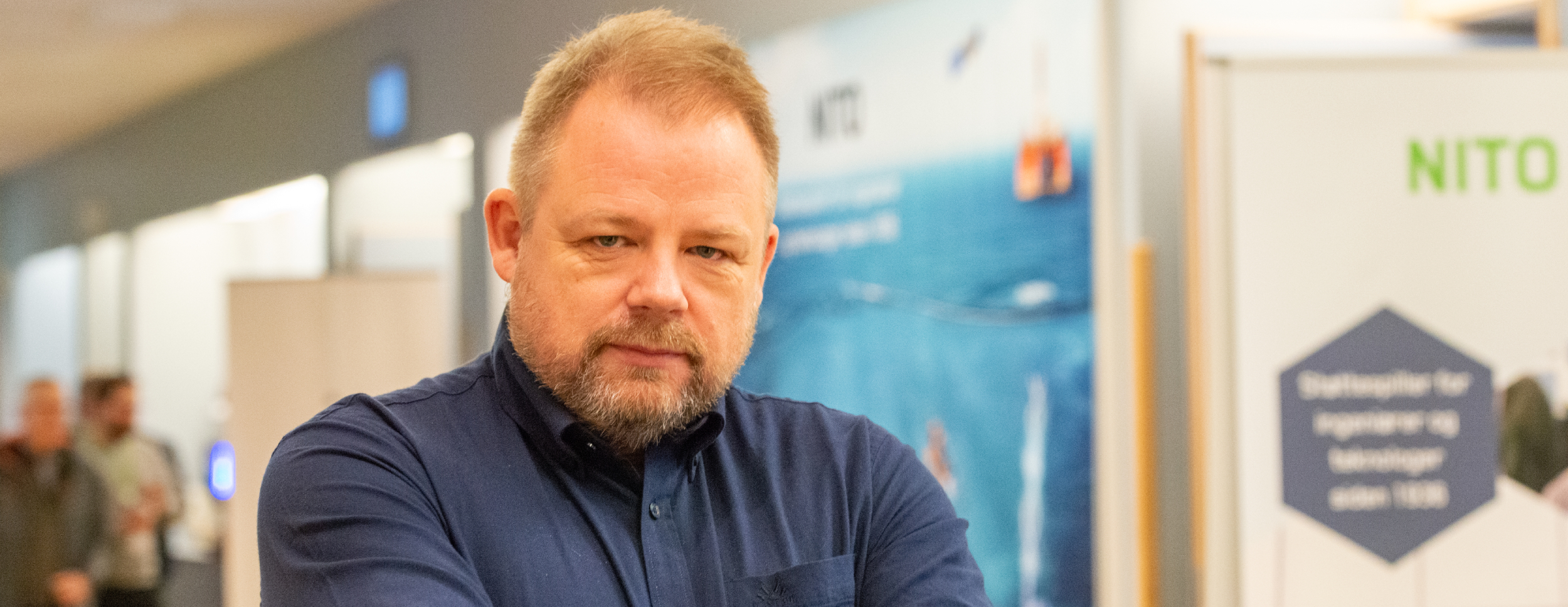 En mann med alvorlig uttrykk står i et moderne messemiljø. NITO-banner i bakgrunnen antyder hans sentrale rolle i organisasjonen.