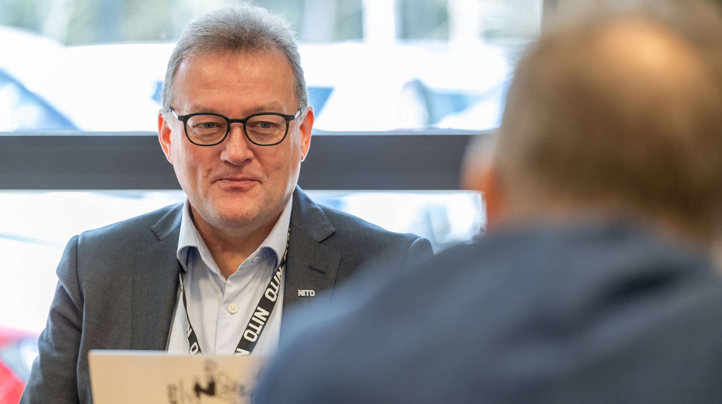 Mann i dress med NITO-navnebrikke sitter i møte. En annen person vises bakfra i et profesjonelt innemiljø.