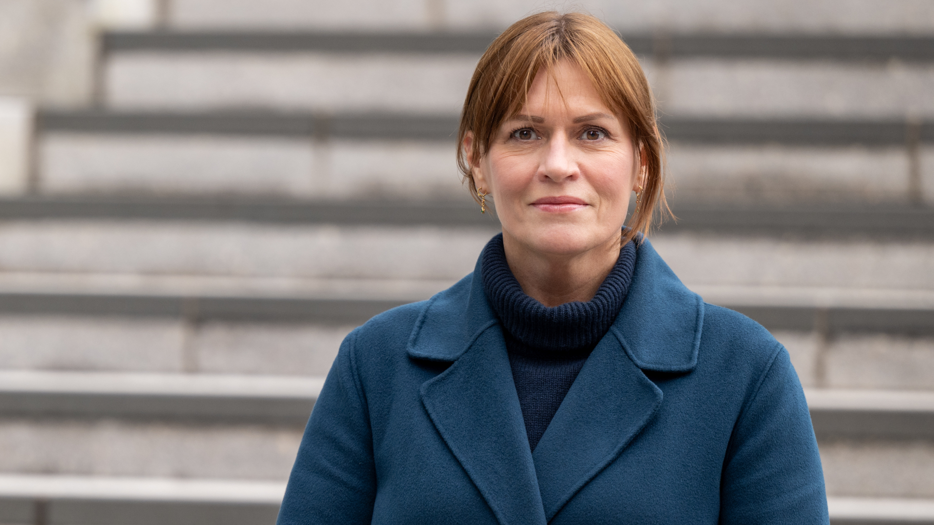Kvinne med høyhalset genser, blå kåpe og brunt hår som ser inn i kamera