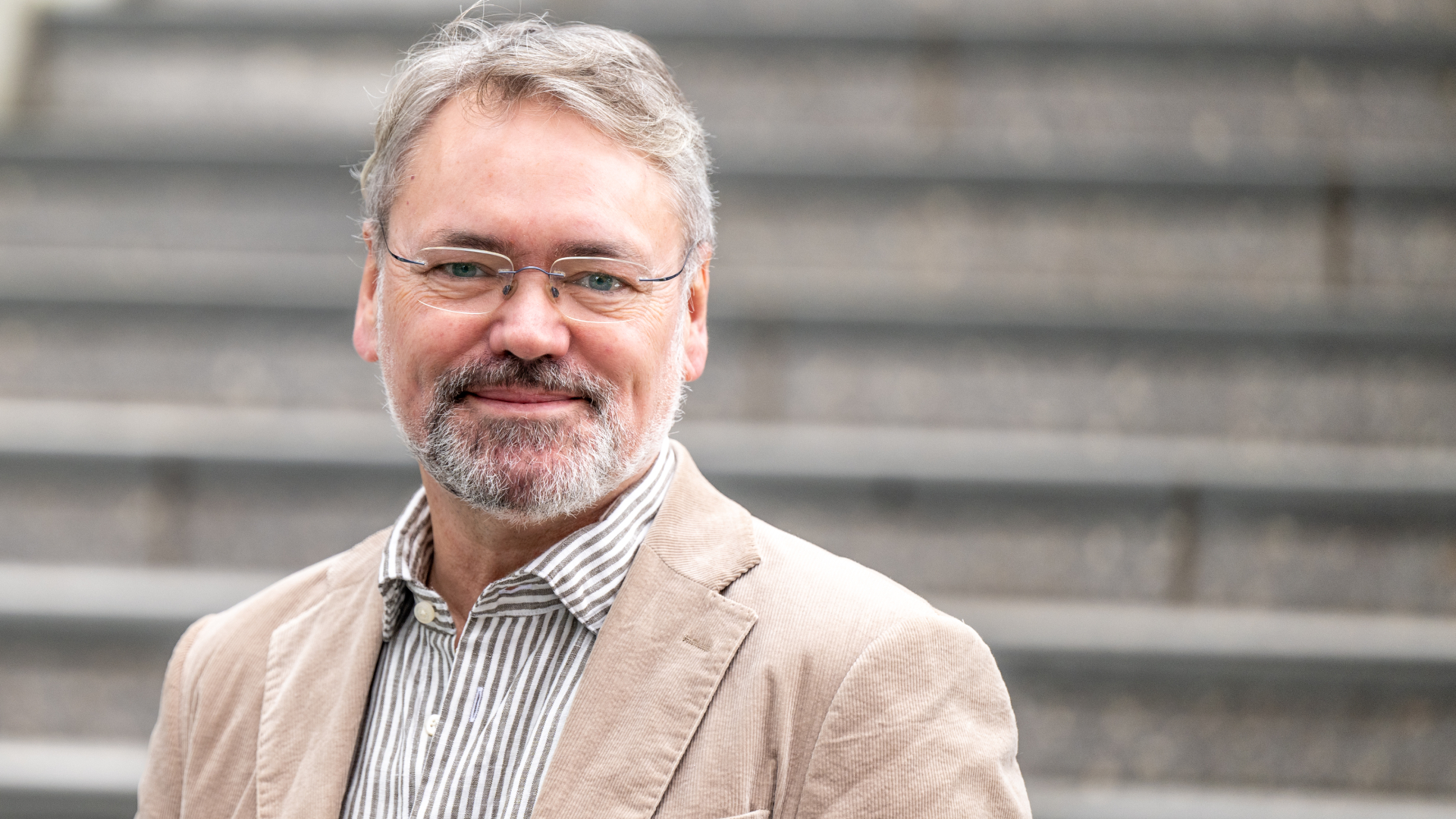 En mann med briller og grått skjegg, med lys blazer og stripete skjorte står foran grå trappetrinn.