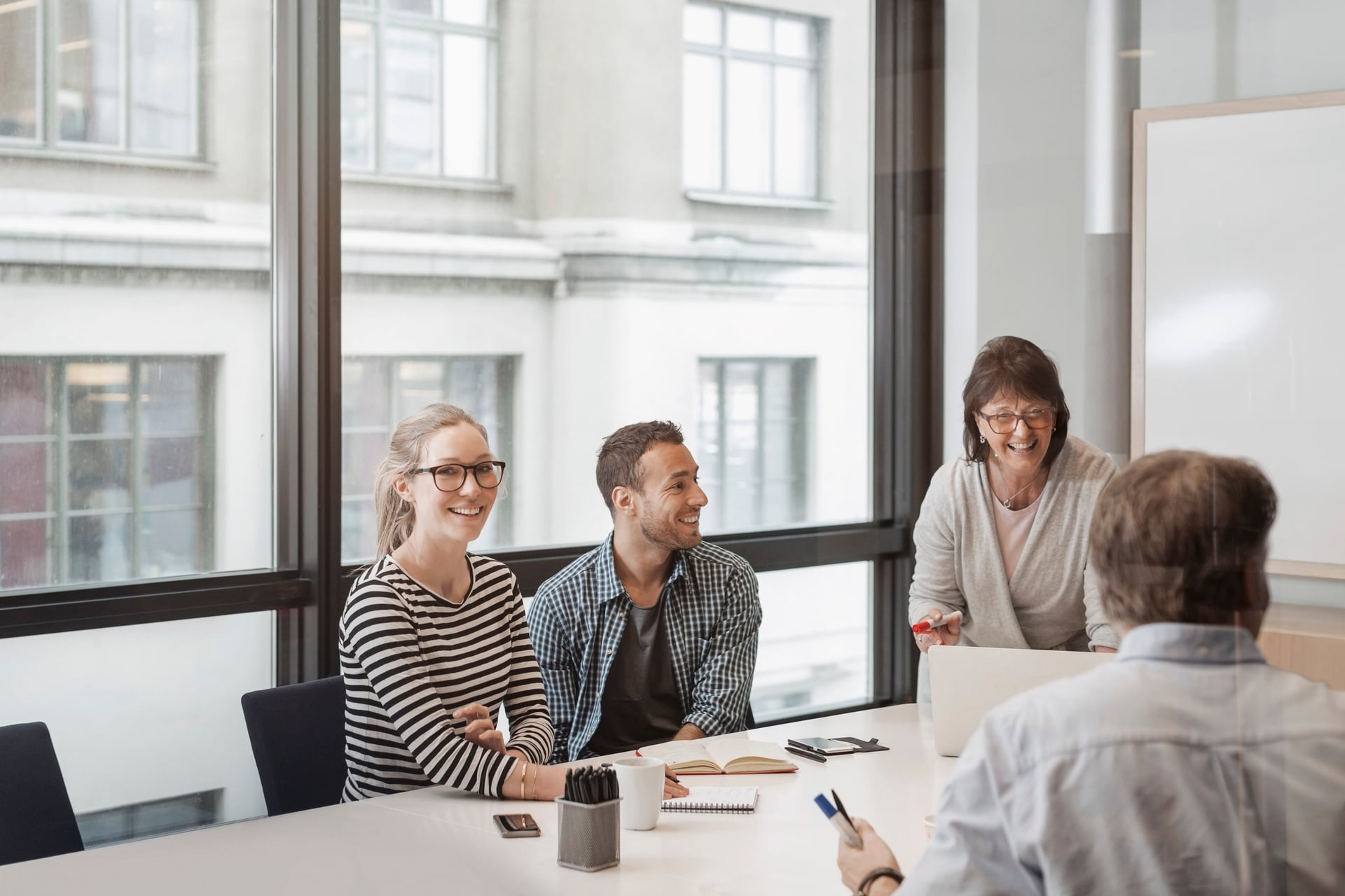 En gruppe kolleger, inkludert en smilende kvinne med briller og en presenterende kvinne, deltar i et positivt forhandlingsmøte på et moderne kontor, som illustrerer samarbeid og dialog i arbeidslivet.