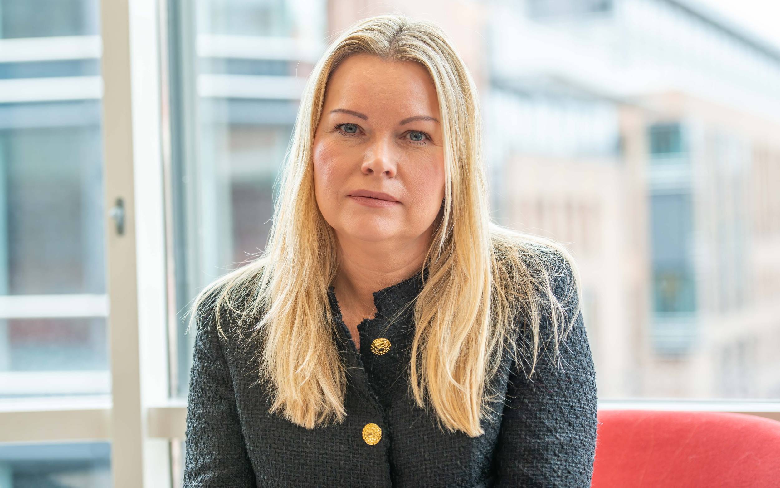 En kvinne med langt blondt hår, iført en mørk jakke med gullknapper, sitter i en lys setting med vinduer bak seg.