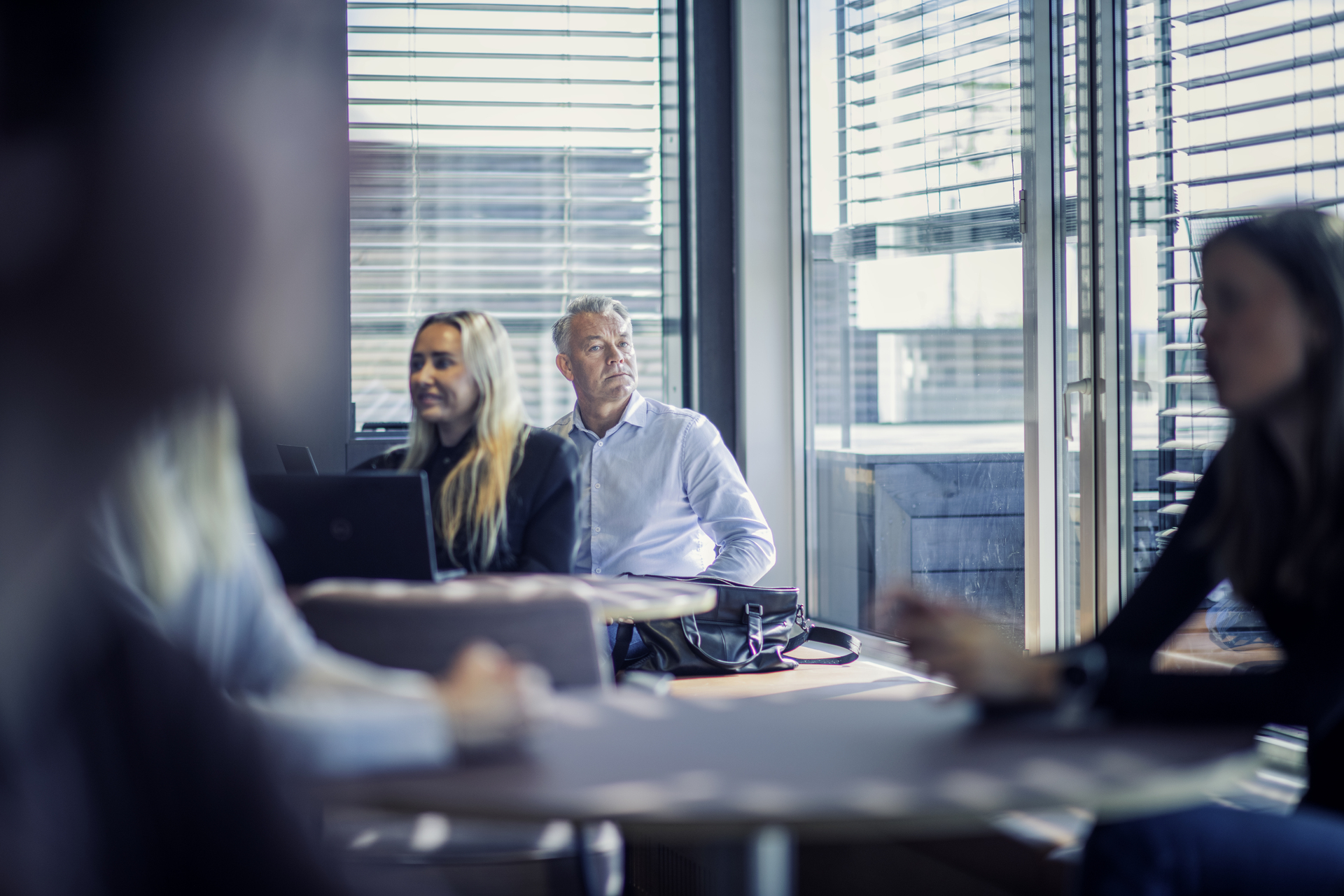 En mannlig ansatt, kanskje en leder, sitter i kontorlokalene på sin arbeidsplass. Rundt han sitter flere kollegaer.