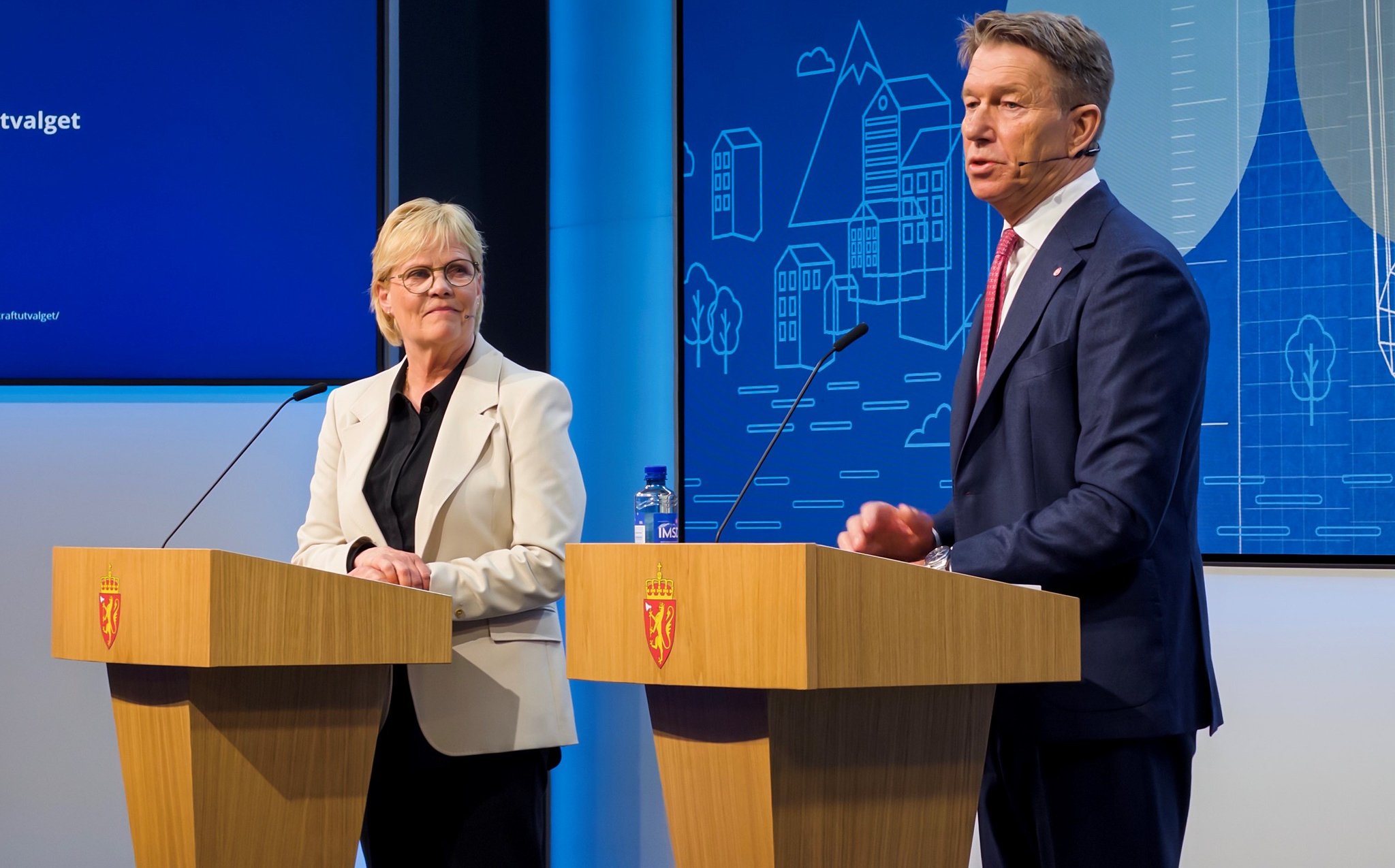 To personer står ved talerstoler på en scene foran en blå bakgrunn med grafikk og norske symboler.