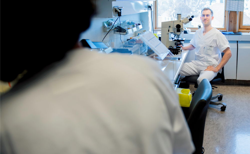 Bildet viser en person i hvit uniform som sitter ved et mikroskop i et laboratoriemiljø. En annen person er i rommet.  