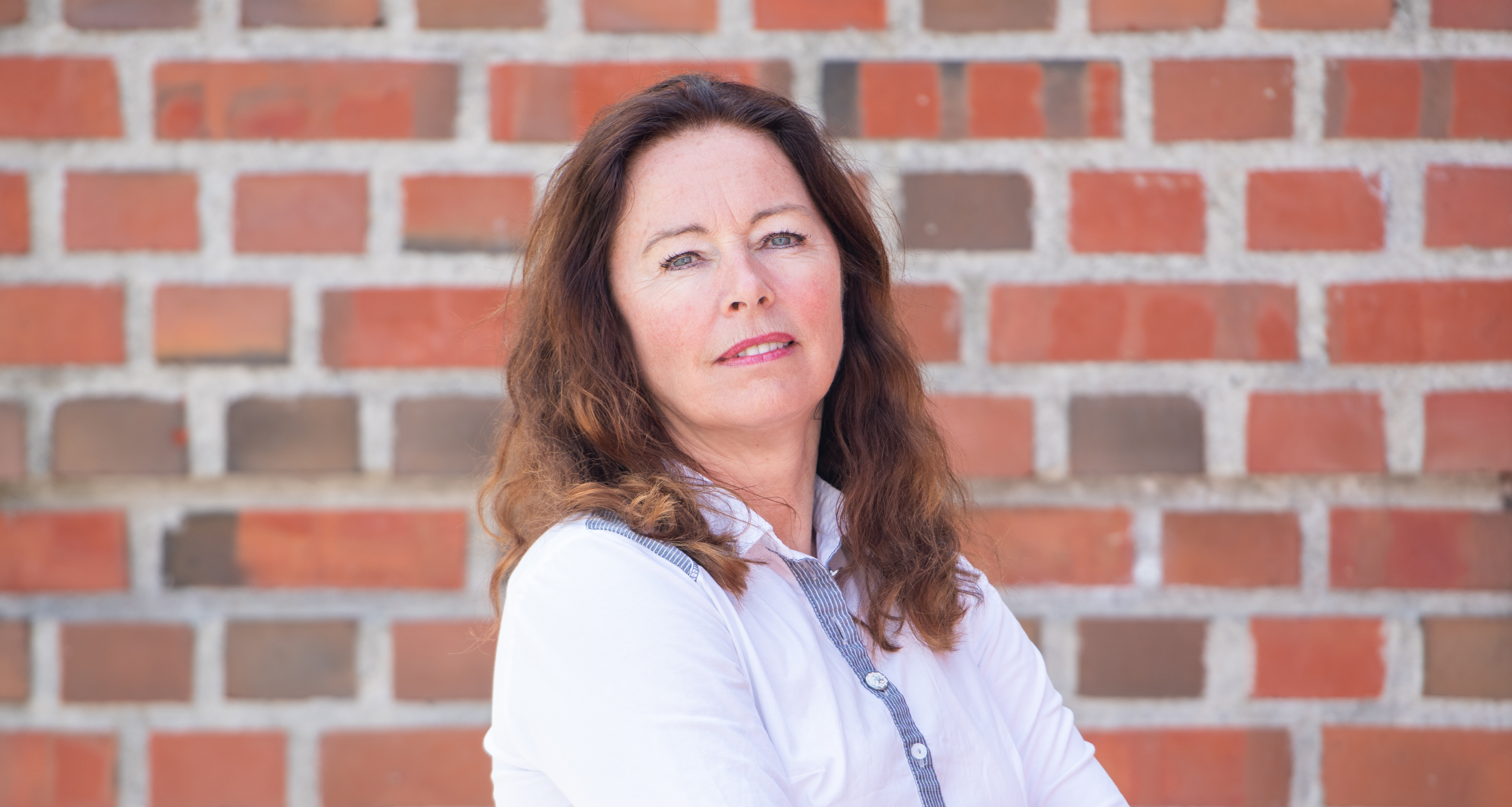 Kvinne i hvit bluse fotografert utendørs foran en teglsteinsvegg.