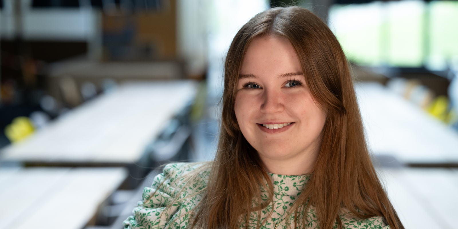 Ung kvinne med langt, brunt hår i grønn og hvit bluse, foran bord og stoler i et rom.