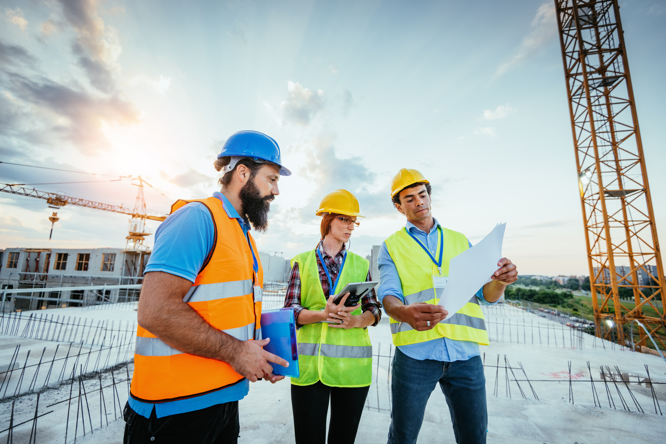 Tre personer med arbeidshjelm og refleksvest studerer tegning på taket av et bygg