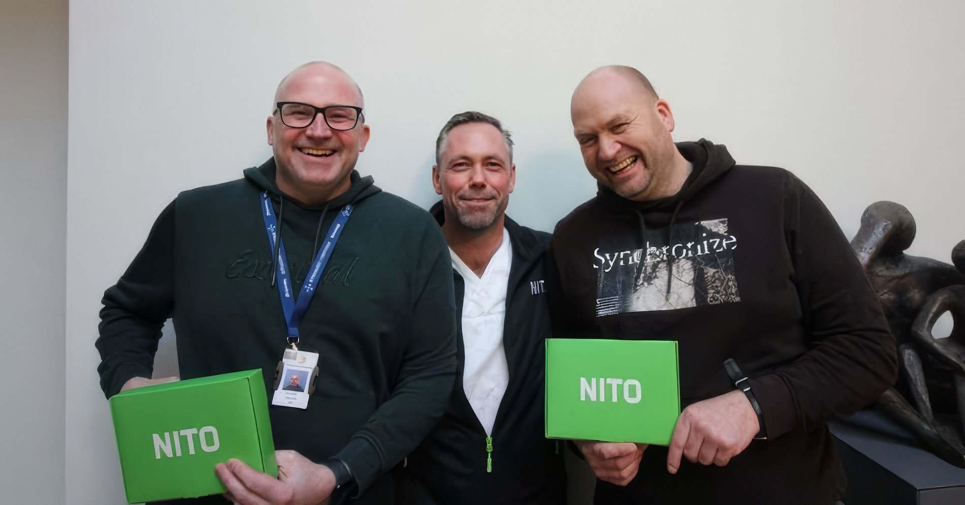 Tre menn smiler bredt. Mannen til venstre har på briller og går i grønn hettehenser, han i midten har på scrubs, og han til høyre har på svart hettegenser