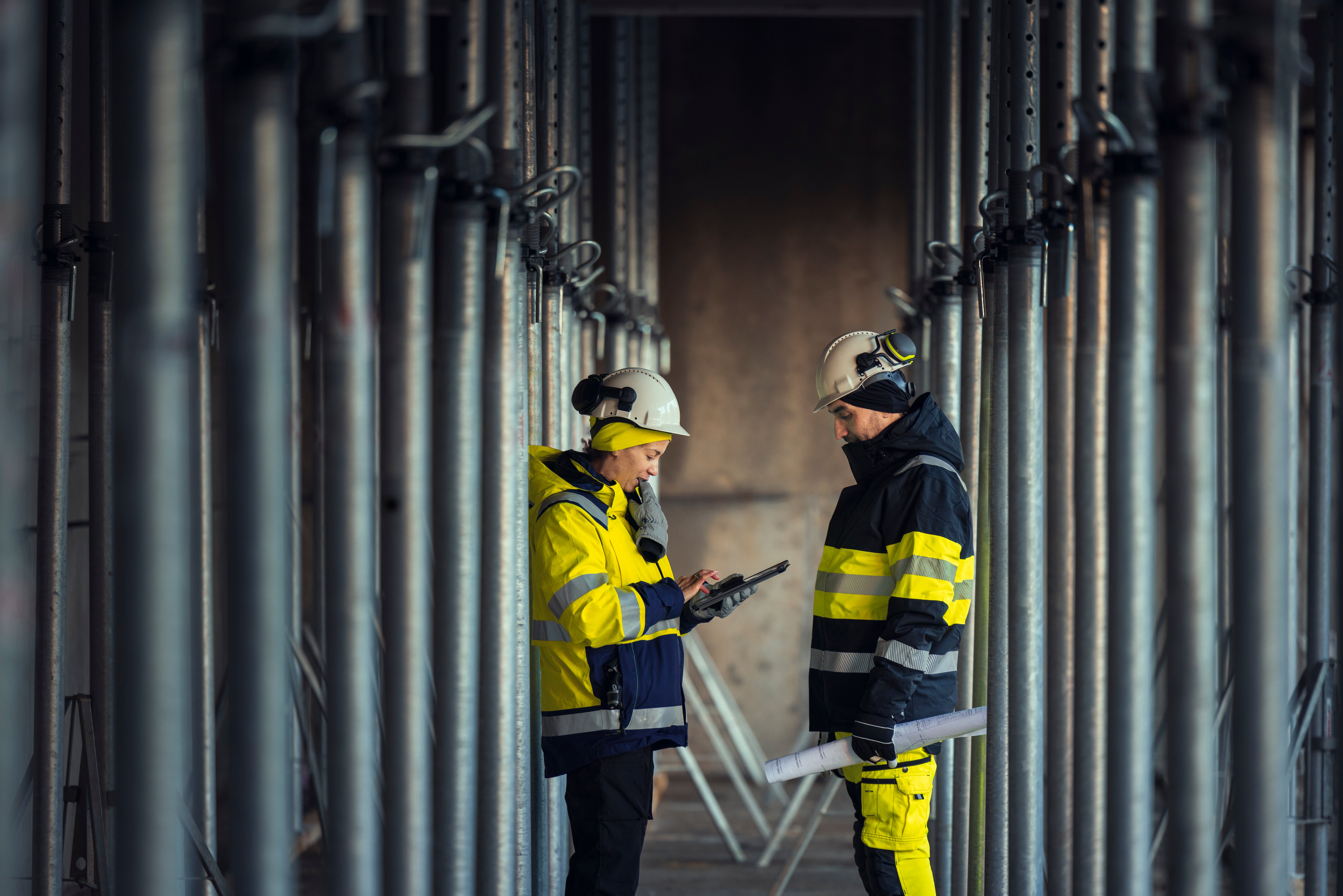  To ingeniører, mann og kvinne, iført arbeidstøy og hjelm snakker sammen i et anlegg med rør i bakgrunnen foto:GettyImages