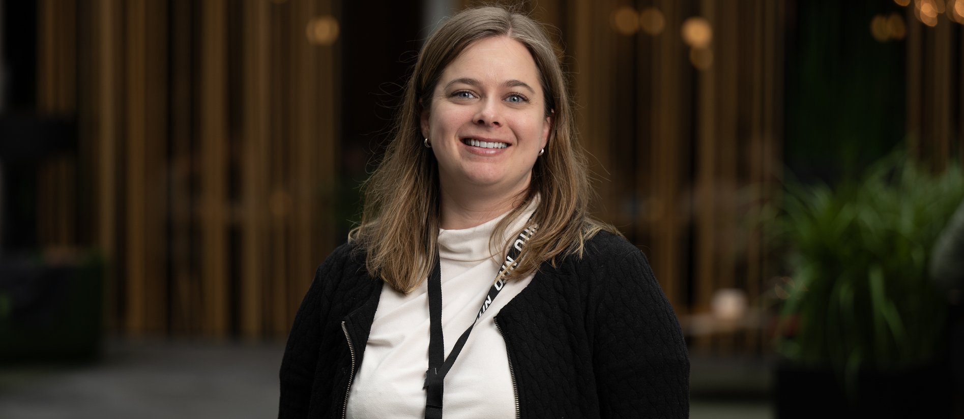 A person with medium-length brown hair stands in front of a background of green plants and light, vertical wooden slats, wearing a light top and dark jacket.