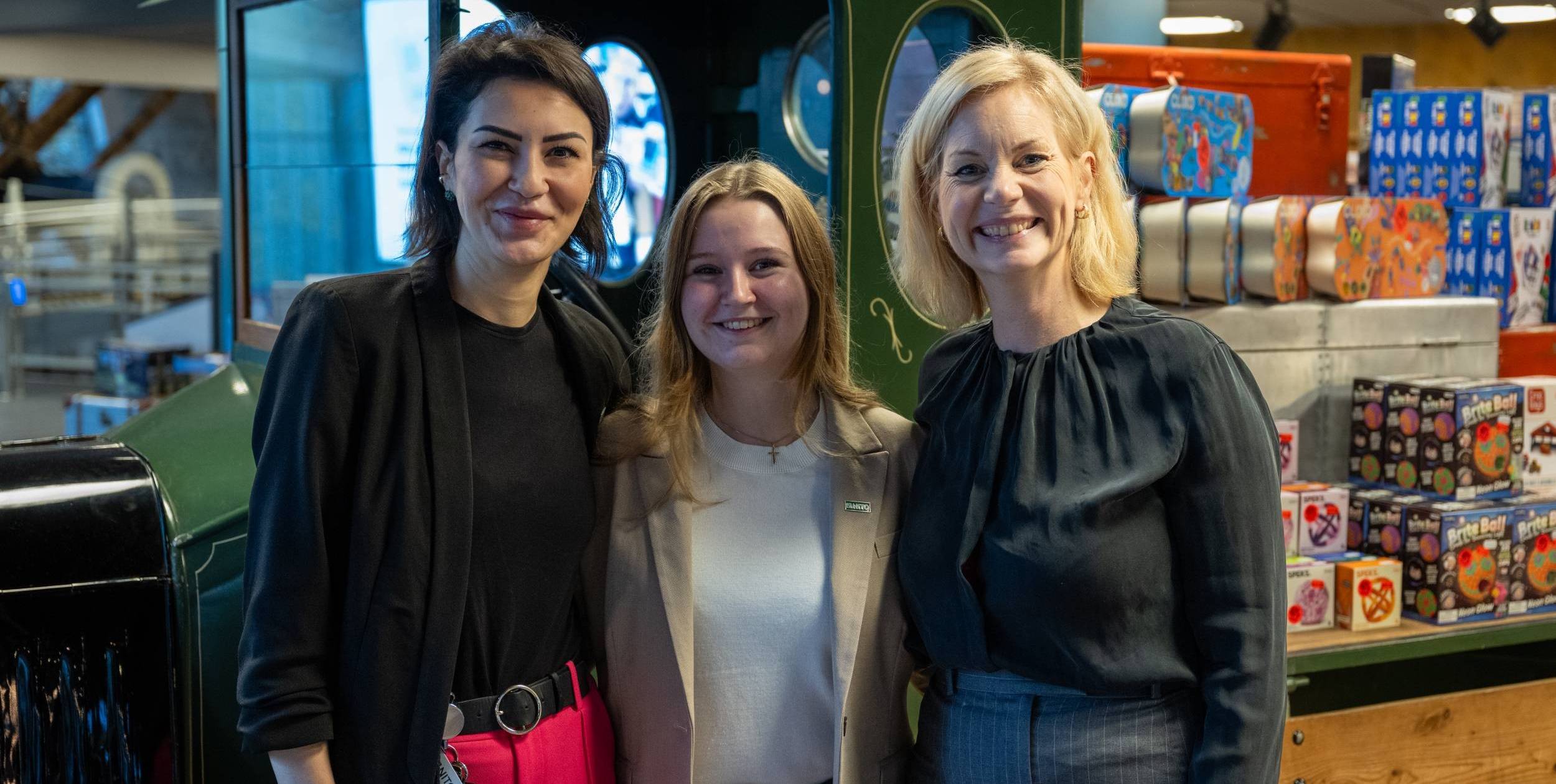 Tre kvinner står tett sammen og smiler, omgitt av fargerike esker og industrielt interiør.