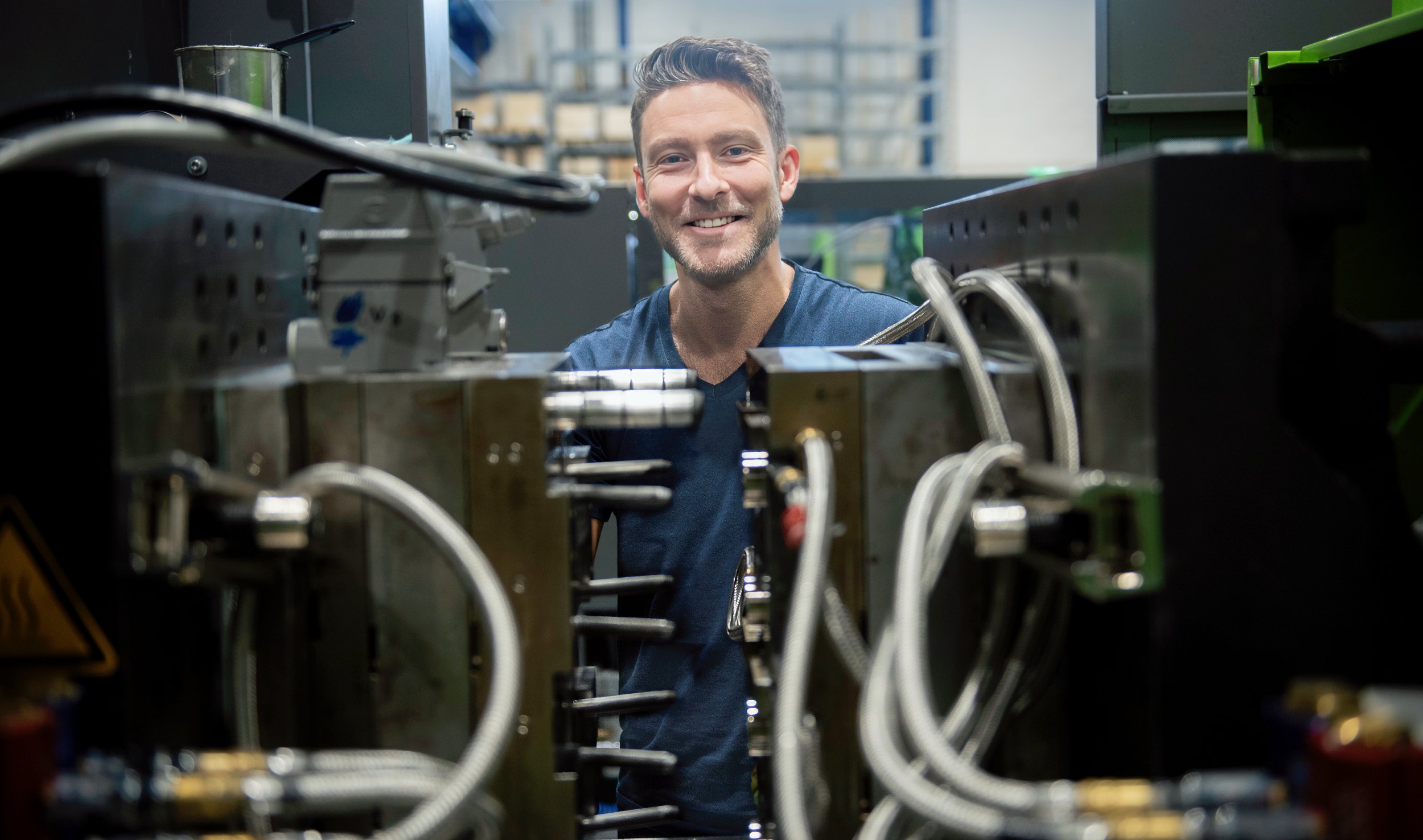 En person arbeider ved en stor industrimaskin omgitt av metallkomponenter og kabler i et produksjonsmiljø.