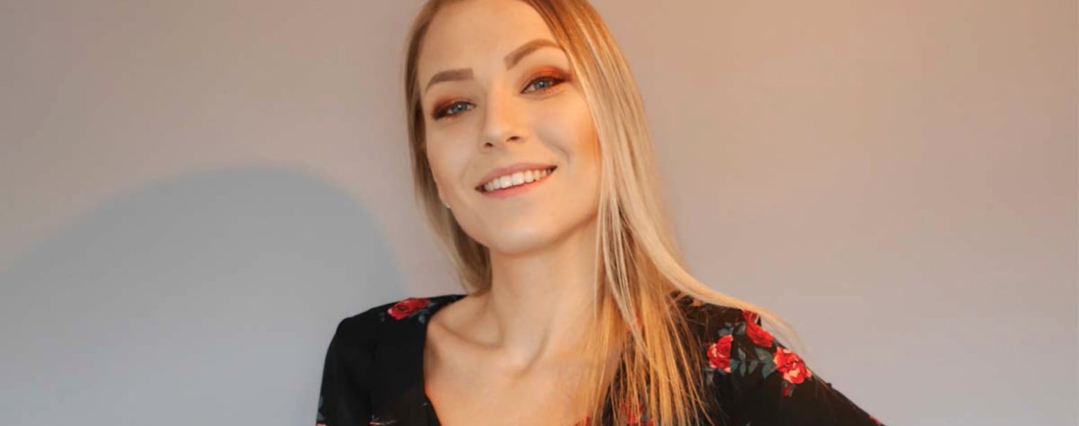 Bildet viser en ung kvinne med langt, lyst hår og blomstrete topp som smiler mot kameraet.