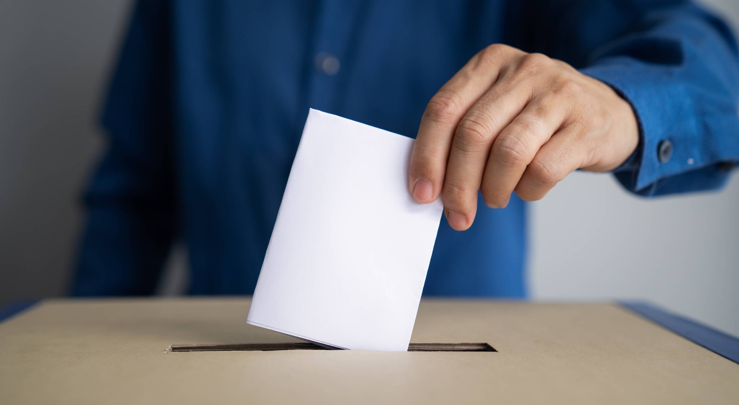 Illustrasjonsfoto av mann med blå skjorte som putter en valgseddel ned i en stemmeboks