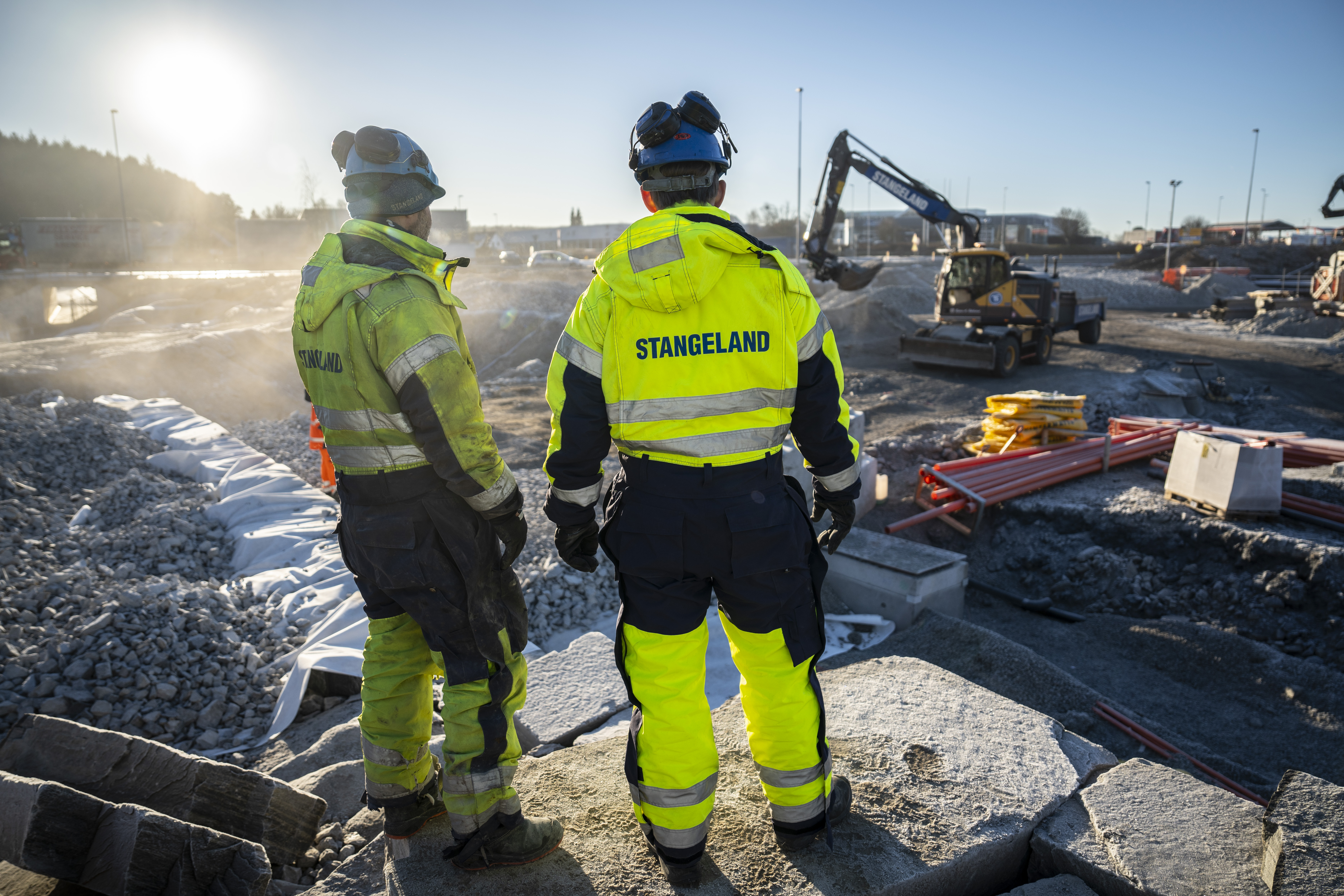 Image of two construction workers looking out at a road project. Excavator and construction work in the background.