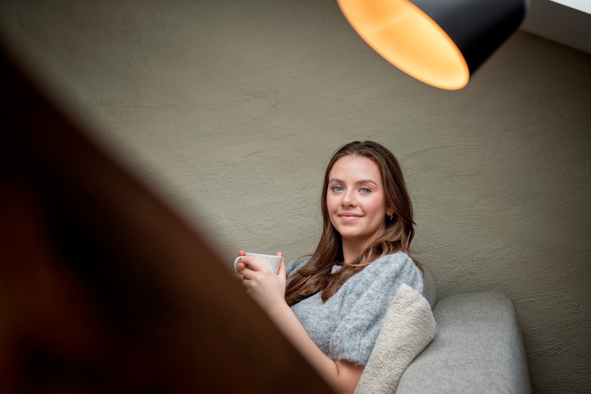 Ung student sitter med kaffekopp i sofa under en lampe. 