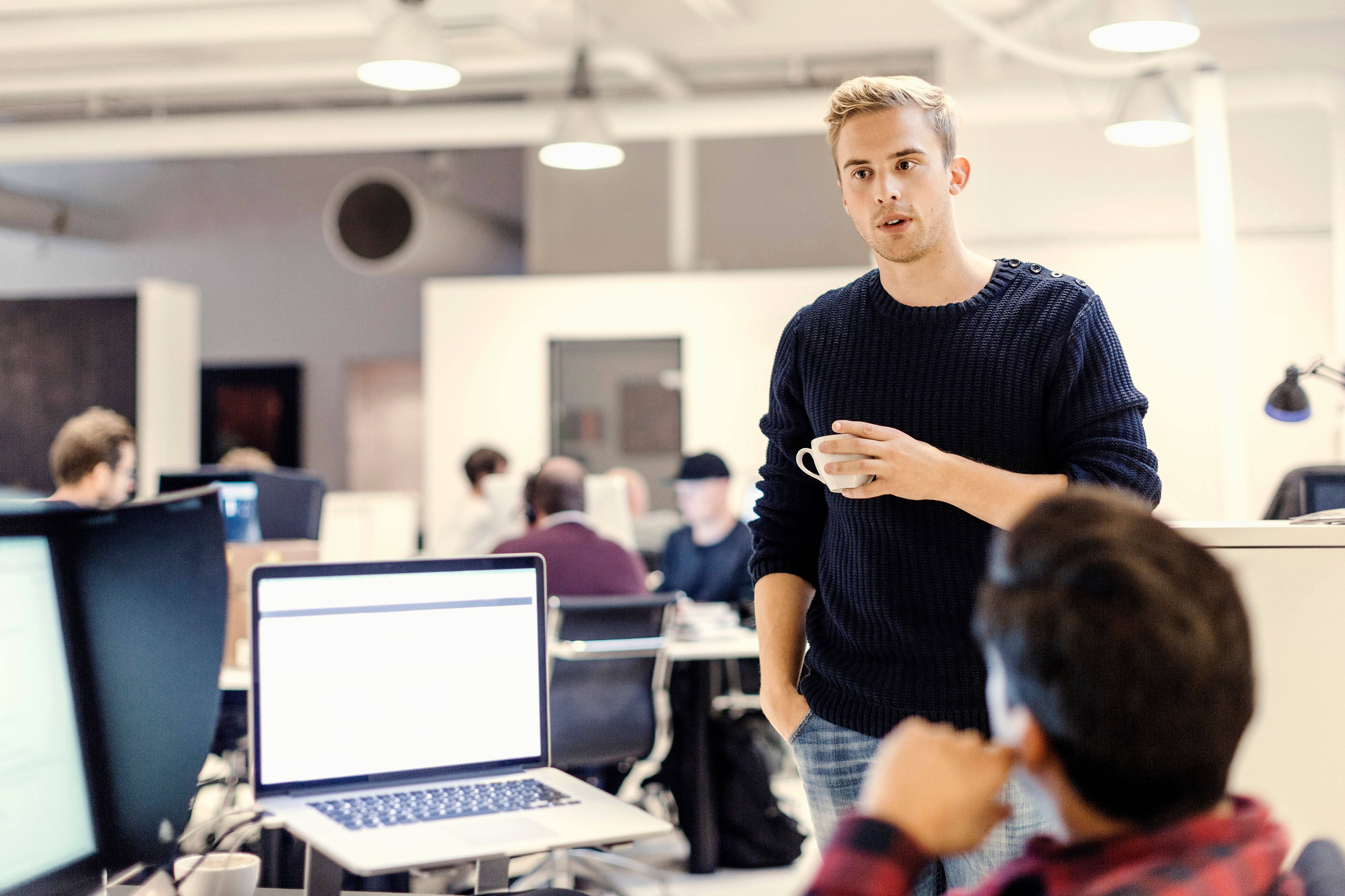 A young male employee stands at a colleague's desk, in an open-plan office, and talks to him. Photo: GettyImages