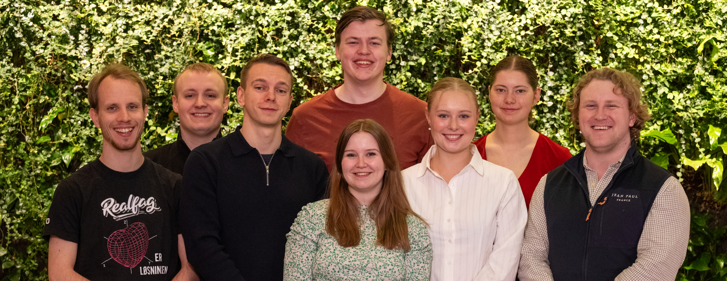 Bildet viser åtte personer foran en vegg av grønne planter, kledd i ulike hverdagsklær.