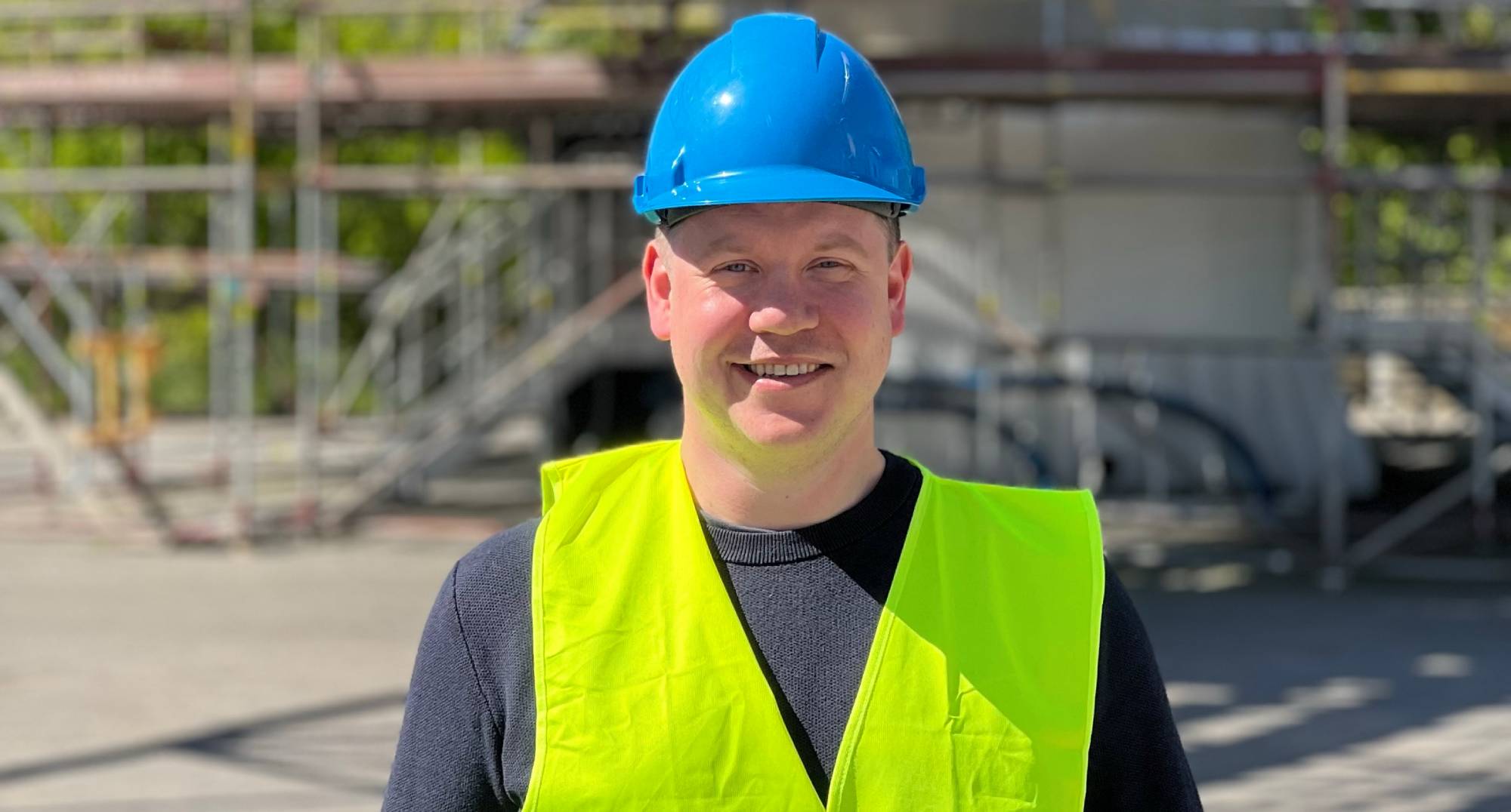Smiling man with a safety vest and blue protective helmet standing on a construction site.