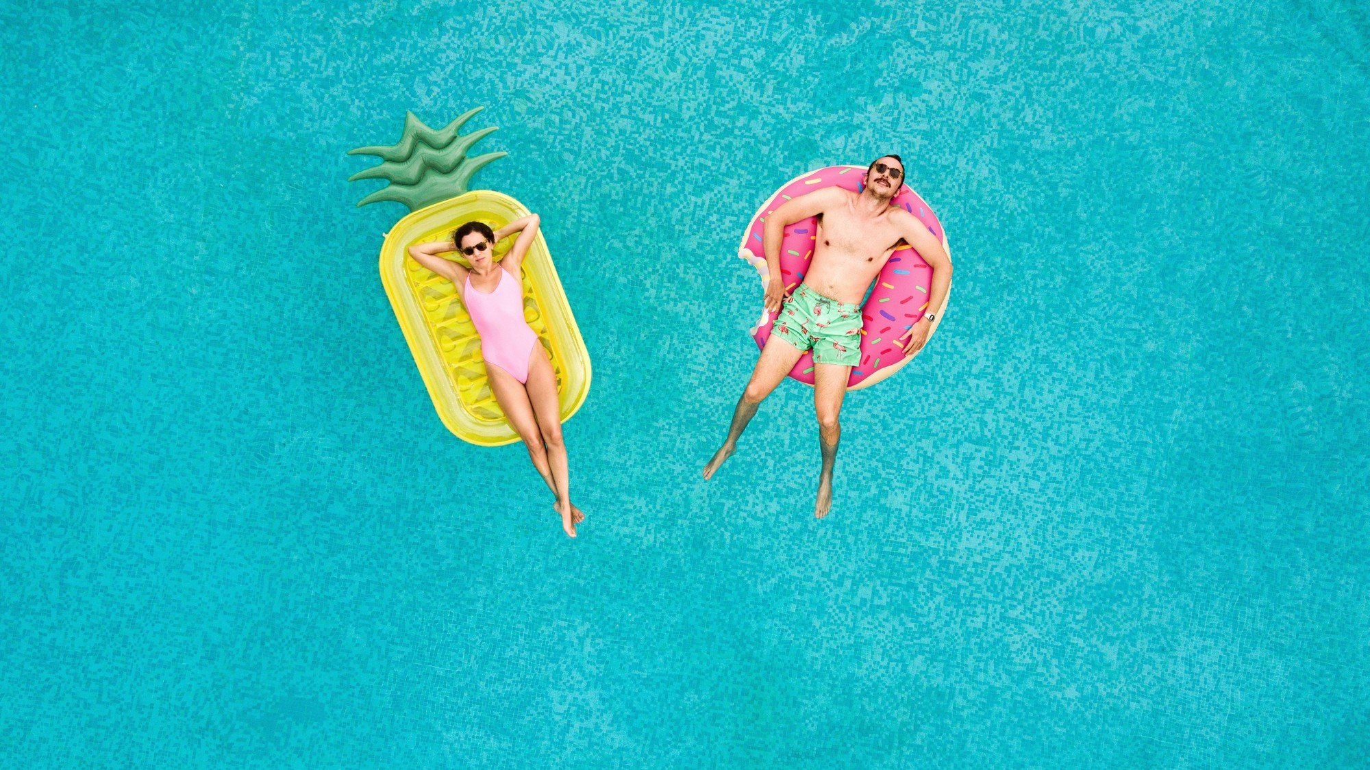 To personer som slapper av på hver sin flytemadrass i basseng 