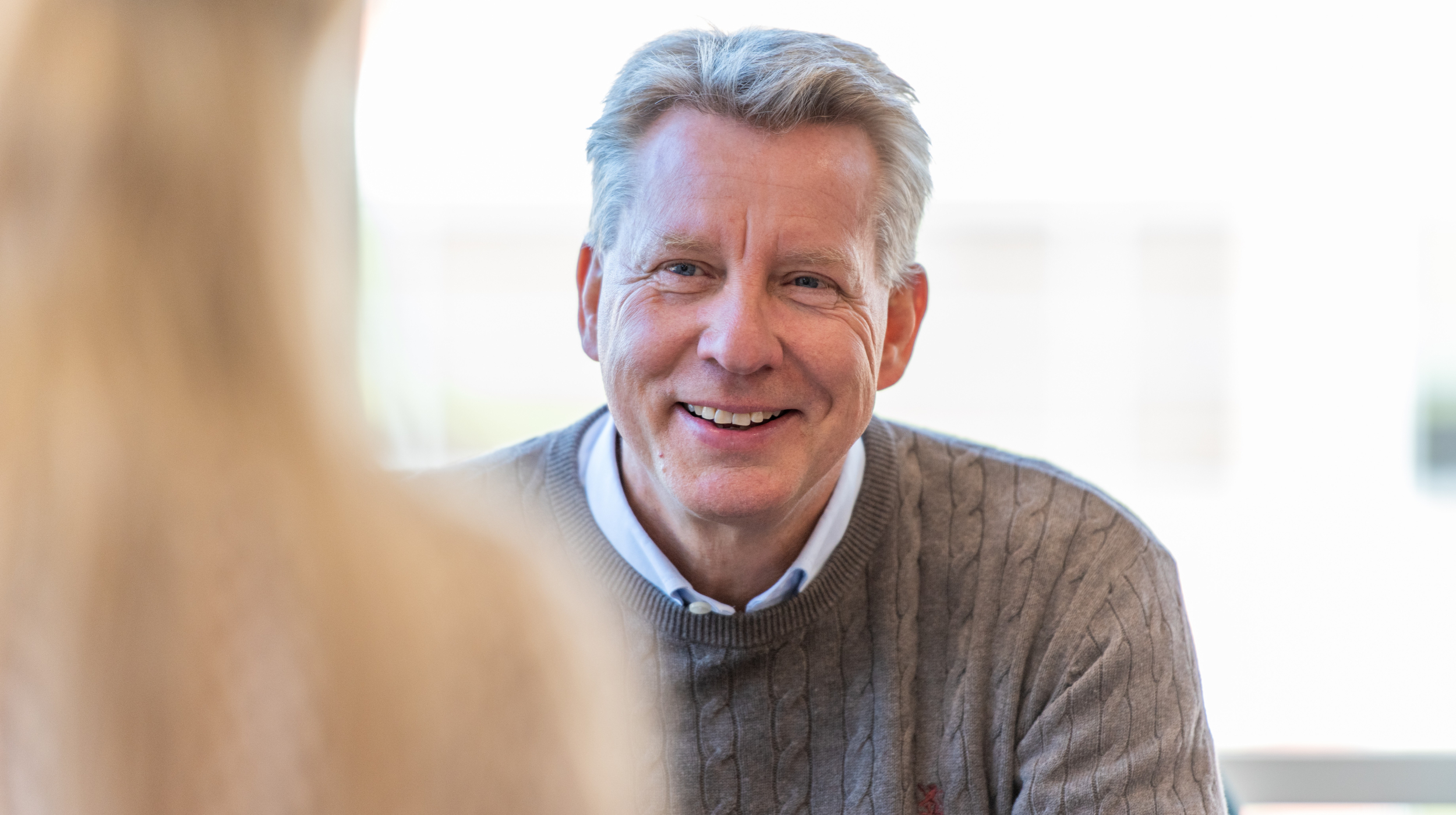 Smilende mann med grått hår, brun genser og blå skjorte. Bakgrunnen er uklar.