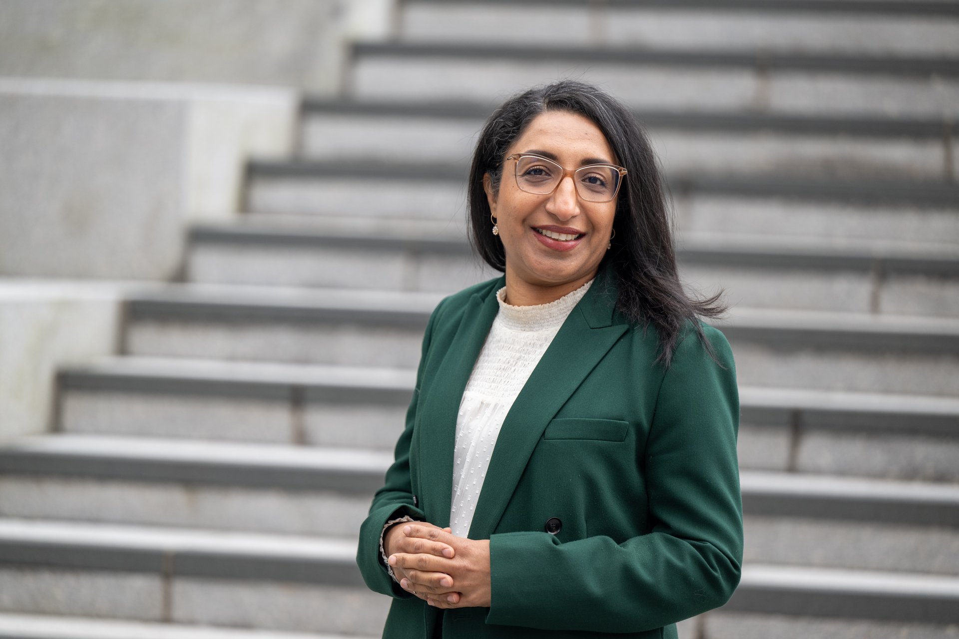 Kvinne med svart hår og briller, har på seg hvit topp og grønn blazer. Står foran en trapp
