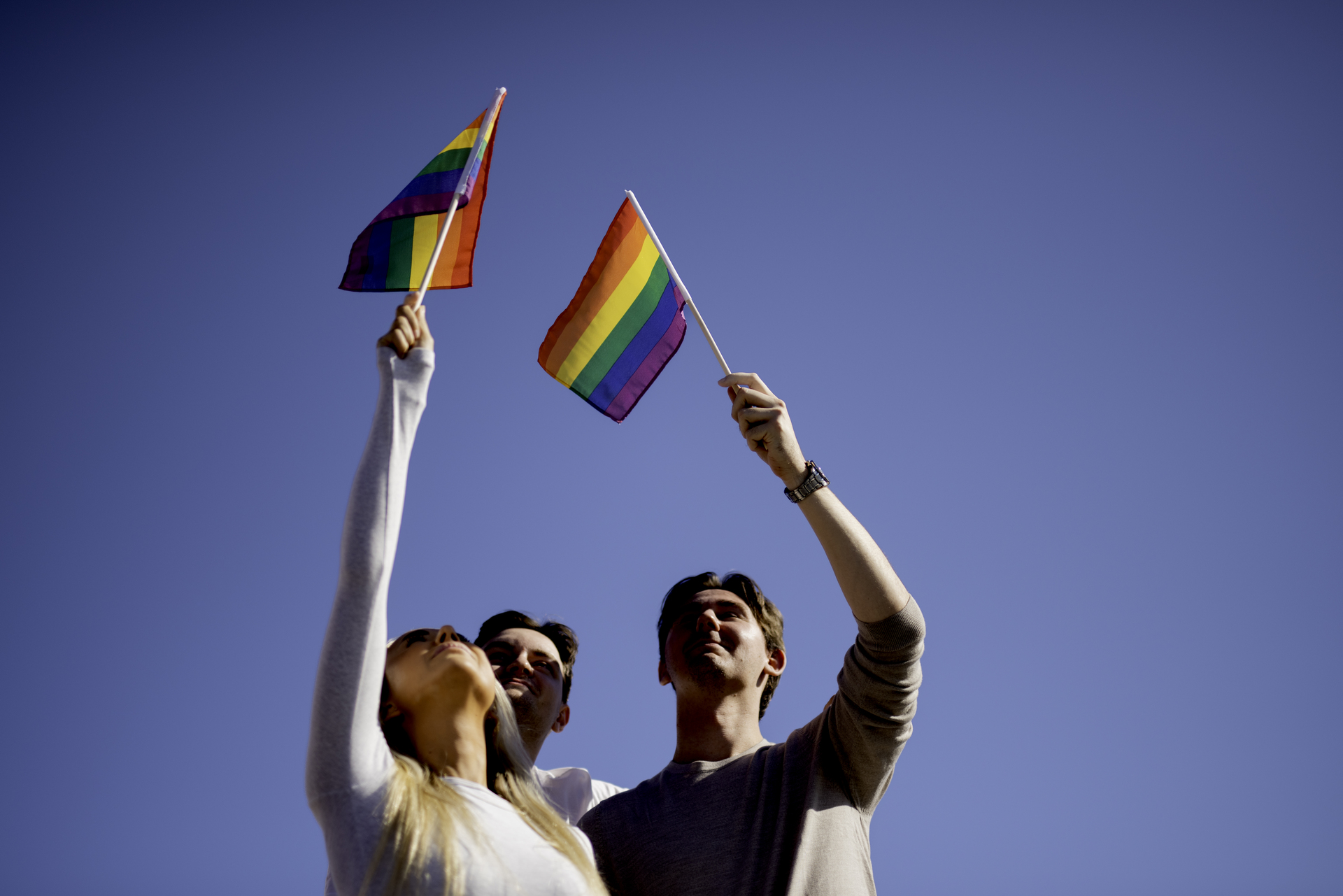 Tre personer holder Prideflagg høyt mot blå himmel