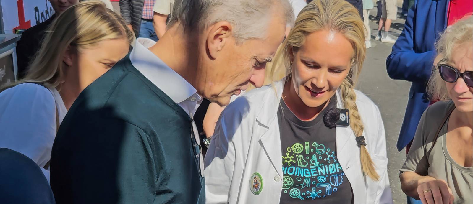 Statsminister Jonas Gahr Støre står sammen med flere personer utendørs, blant annet en kvinne i hvit frakk og en sort t-skjorte med teksten "Bioingeniør"