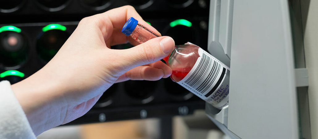A hand places a small glass bottle of red liquid in a machine. The bottle has a label with a barcode. The background is technological.