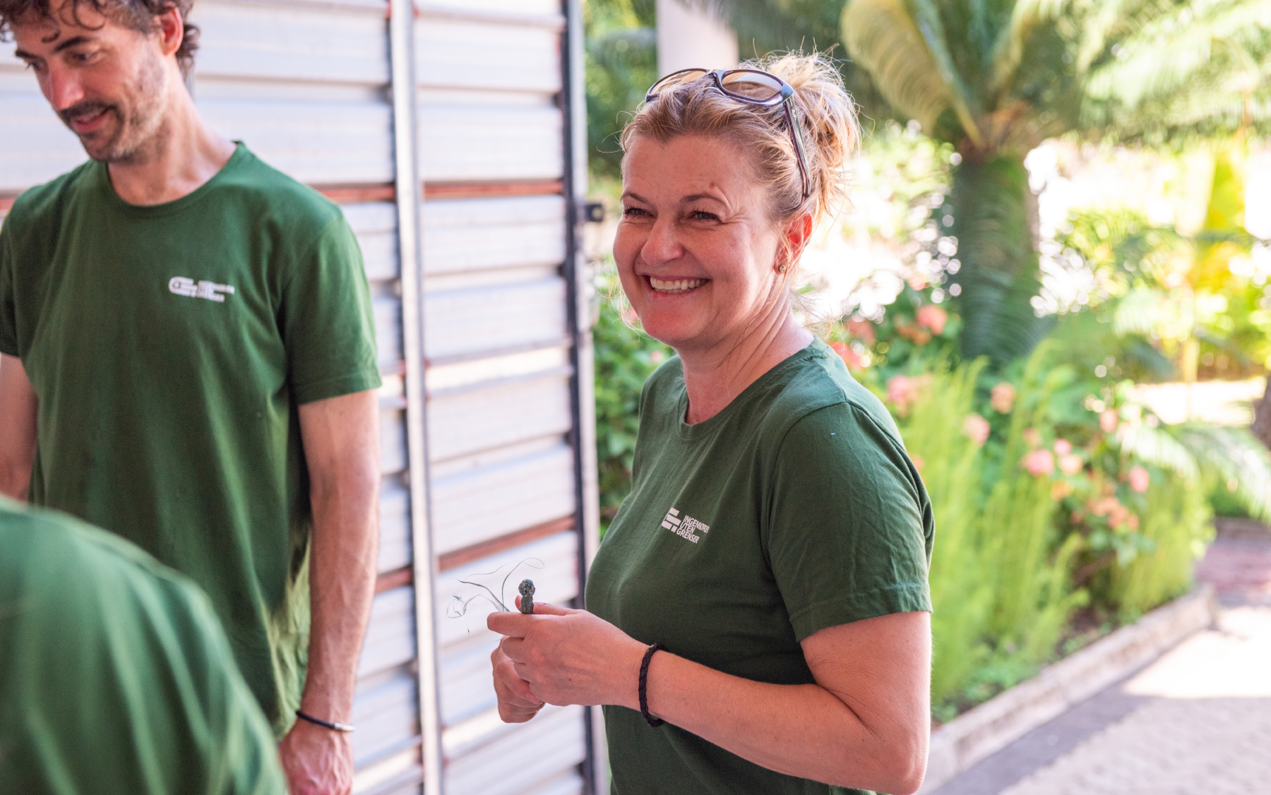 To personer i grønne t-skjorter står foran planter og en garasjeport. Stemningen er avslappet.