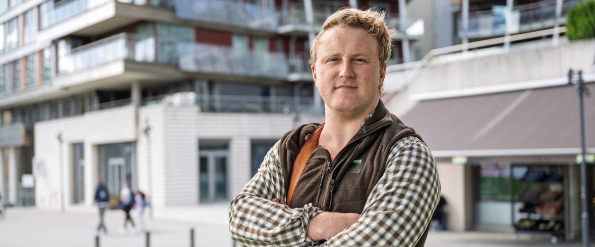 Jaradn Byrkjeland i rutete skjorte og vest står med armer i kors foran bygninger. 