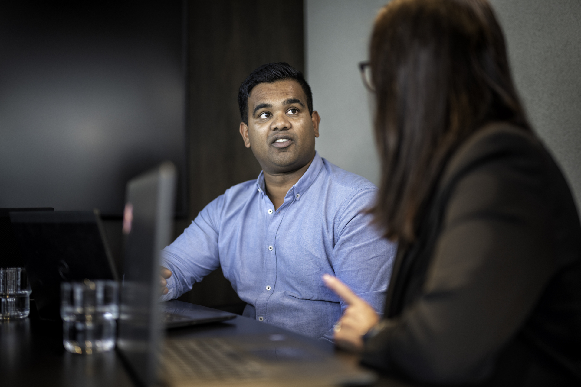 Two people are sitting at a table with a computer on the table. A man in a blue shirt and a lady who you can see from behind.