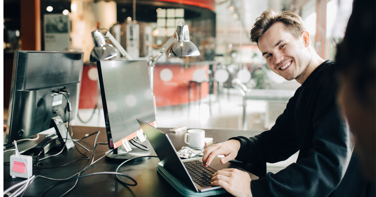 A young male employee sits at his desk in a modern open-plan office, smiling at the camera while working on his computer.