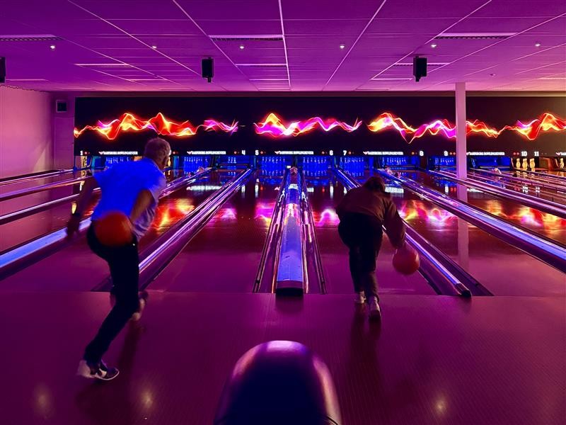To personer som er i gang med å kaste bowlingkula fremover mot kjeglene. Lilla lys i rommet. Kredit: Nathalie Oberti Karlsen