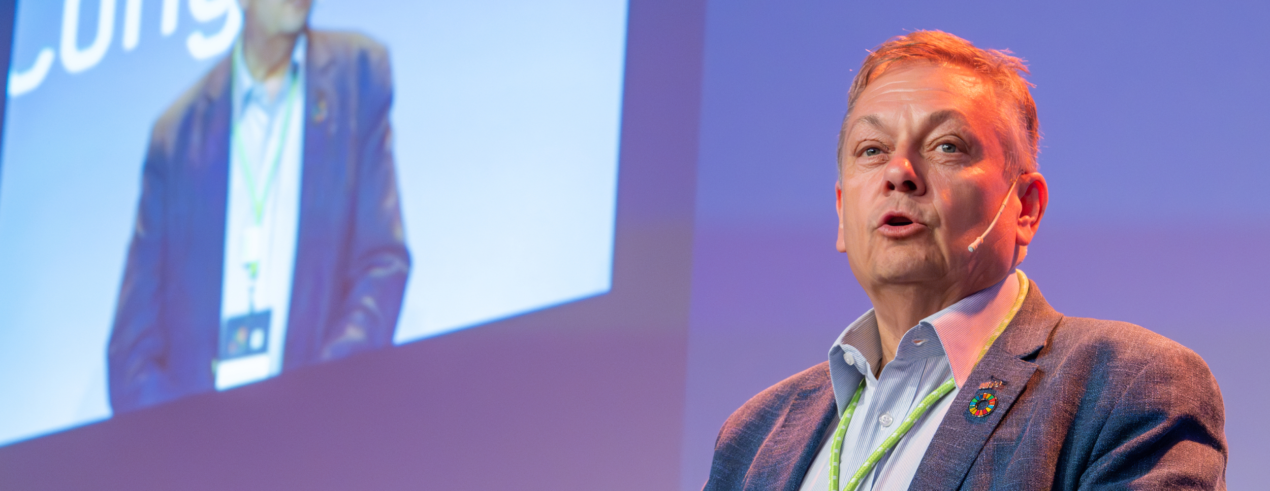 A person in a suit with a UN pin stands in front of a large screen showing an enlarged version of himself. The background is blue and professional.
