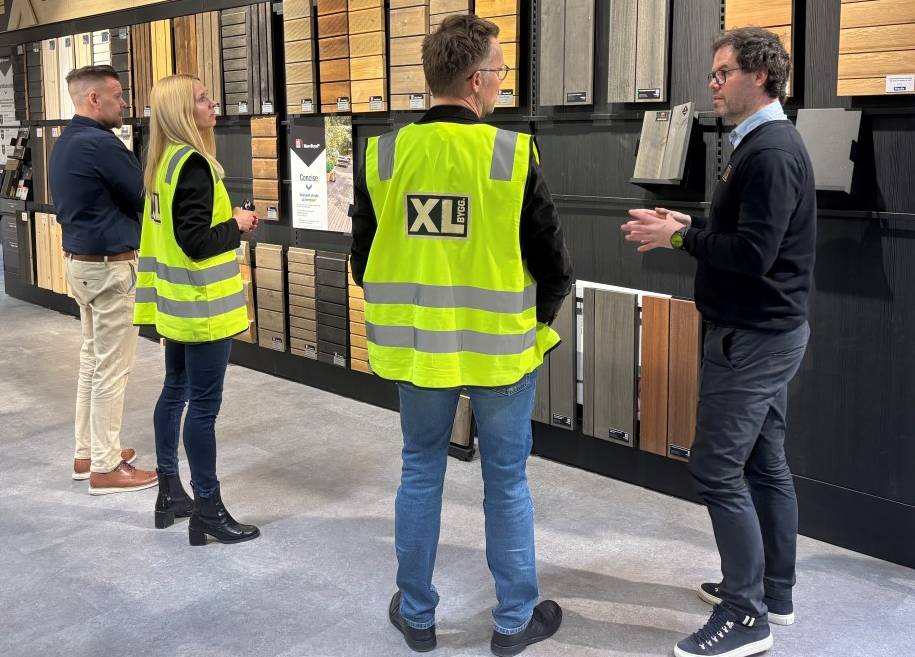 Fire personer i en butikk, to med gule vester, ser på utstilte trepaneler på en vegg. En diskusjon pågår mellom dem.