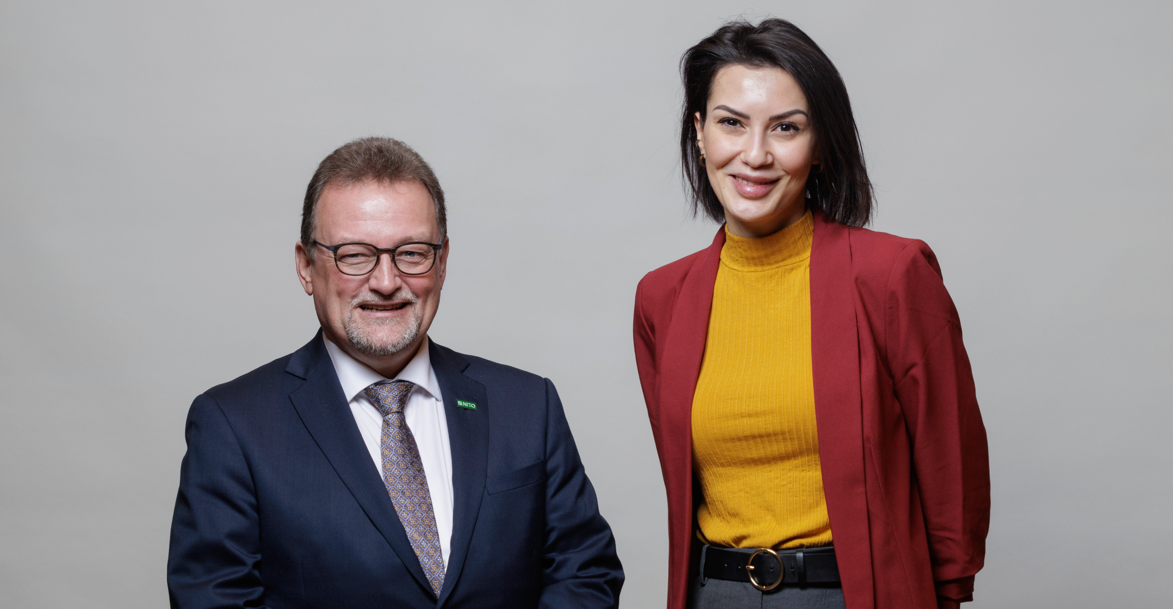 To personer står side om side og smiler. En mann i dress og briller, og en kvinne i rød blazer og gul genser foran grå bakgrunn.