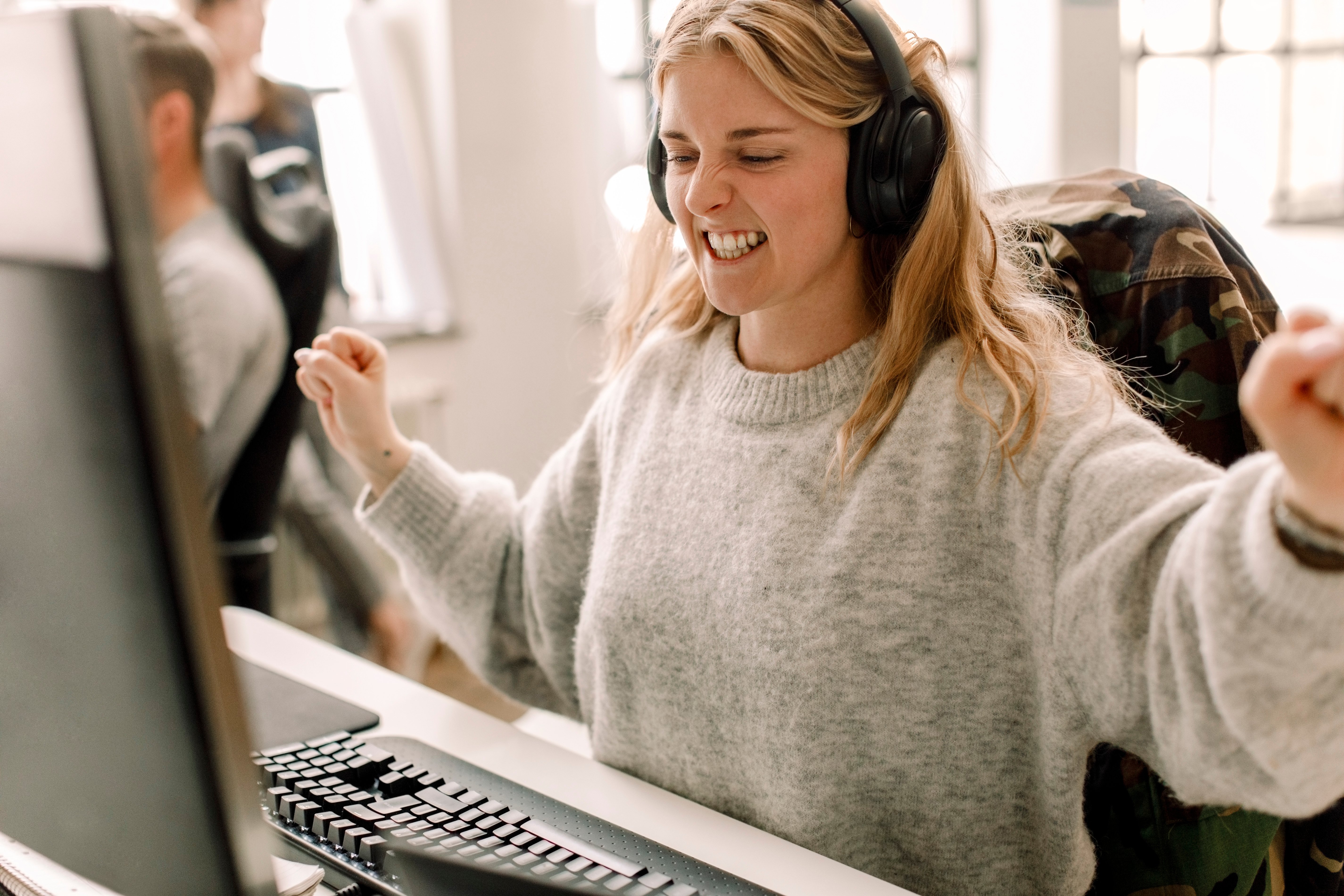 Ung blond kvinne med headset foran en pc-skjerm, hun jubler 