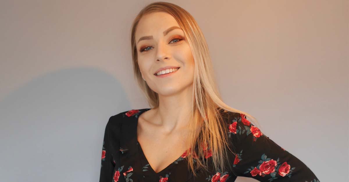 Bildet viser en ung kvinne med langt, lyst hår og blomstrete topp som smiler mot kameraet.