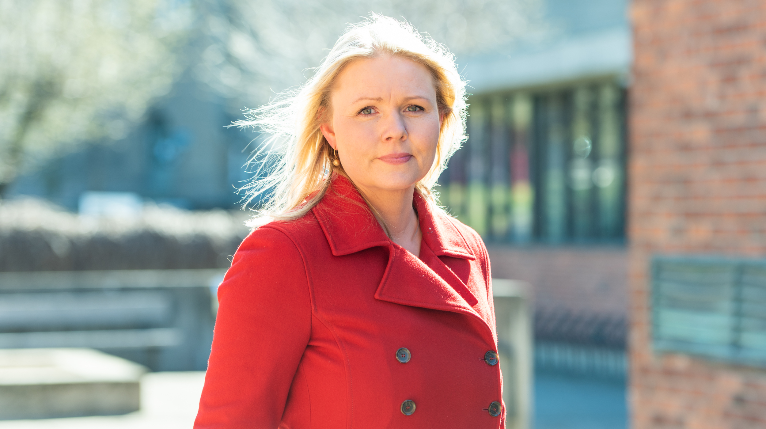 Bilde av kvinne med rød kåpe og lyst hår som ser inn i kamera. Bakgrunnen er uklar, men man skimter bygninger og vinduer. 